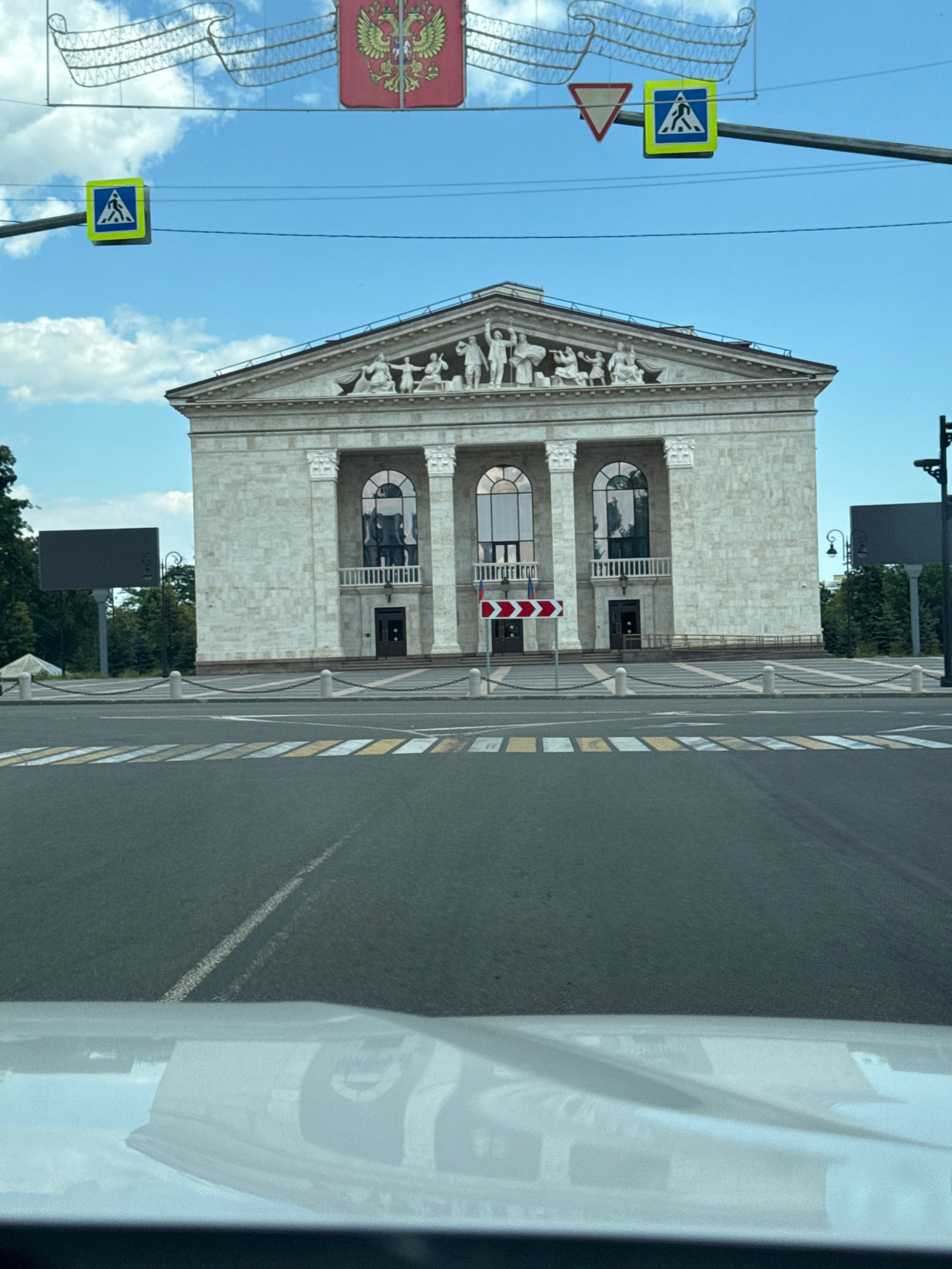 The image depicts a building with a classical facade, featuring architectural reliefs along the pediment. The building is made of light-colored stone blocks and has several arched windows. A road with yellow and white markings runs alongside the building. Several traffic signs are visible, including a pedestrian crossing sign and a warning sign with a red triangle. A large sign with the Russian coat of arms is also present. The sky is blue with some clouds, suggesting daytime. The overall scene appears to be an urban environment, possibly in Mariupol, Ukraine, given the location context. The image is oriented vertically, and a white car's reflection is partially visible on the right side. No specific activity or event is clearly depicted, but the presence of the road and signs implies a public space. The weather appears to be clear and sunny.