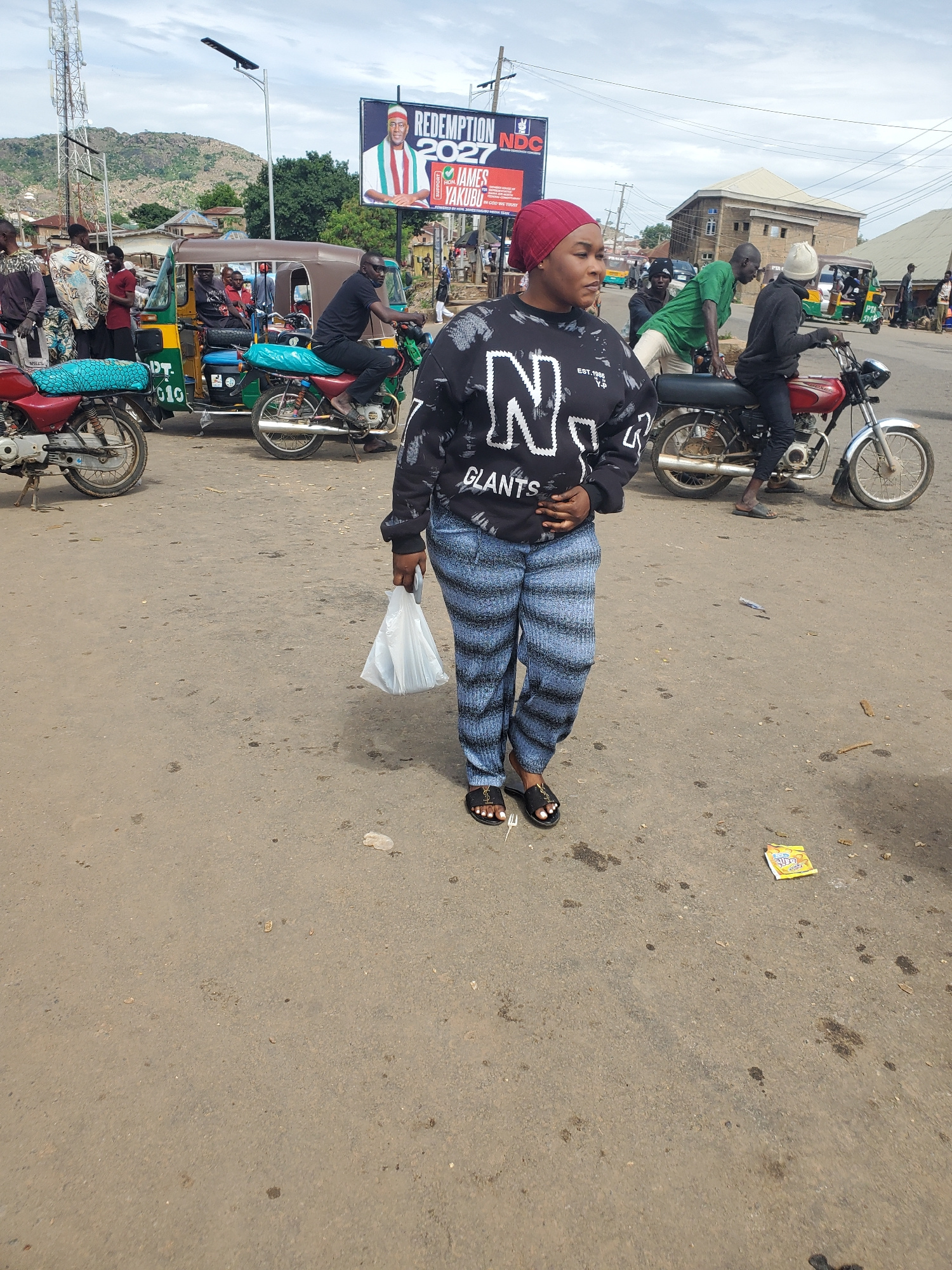 The image depicts a street scene in Gwong, Nigeria, under a cloudy sky. In the foreground, a woman wearing a red headscarf, a black sweatshirt with white lettering, and patterned blue and black pants, walks across the asphalt. She is holding a white plastic bag in her right hand and wears black sandals. To her left, a red motorcycle is parked. Behind her, a green and yellow auto-rickshaw with passengers is visible, and a man is riding a red motorcycle. Further in the background, a large billboard with an image of a man and the text "REDEMPTION 2027 NDC JAMES YAKUBU" is displayed. Several other people and motorcycles are scattered throughout the scene, and the environment appears to be a busy marketplace or transportation hub.