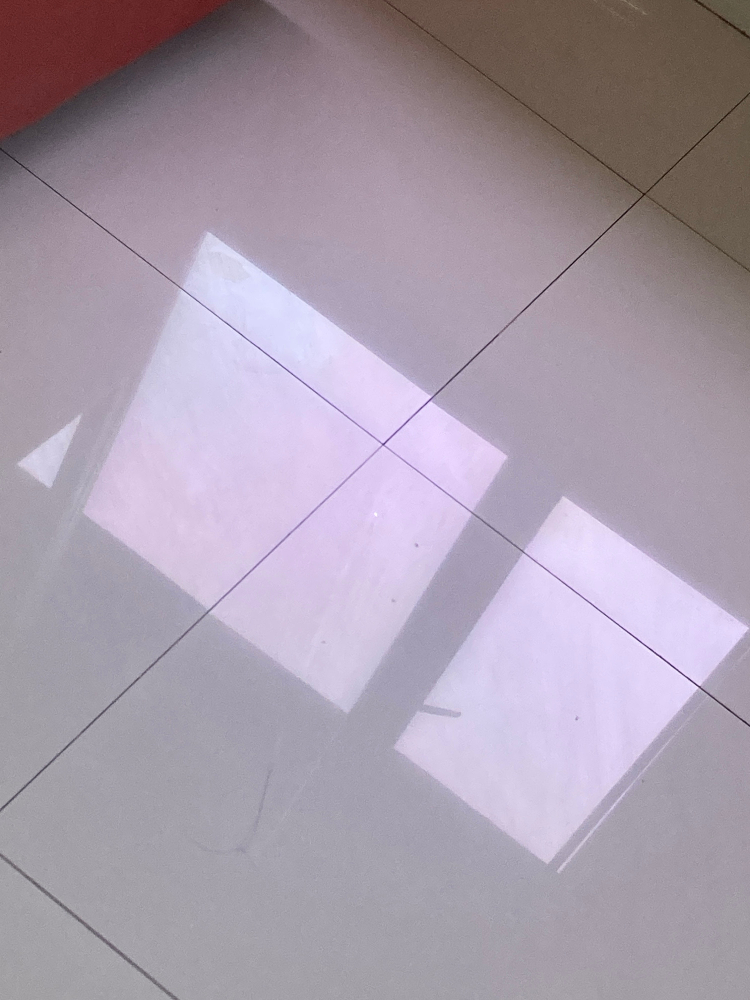 The image shows a tiled floor with two rectangular reflections of light. The tiles are square and arranged in a grid pattern, with thin dark grout lines. The reflections are bright and pinkish-white, suggesting they are from a light source such as a window or a lamp. There is also a small triangular reflection in the upper left quadrant. The upper portion of the image features a blurry red object, possibly furniture or a wall. The floor appears to be clean. There are no people or animals visible in the image. The image is taken from a high-angle perspective, looking down at the floor. The location is stated to be in the city of Proyonangg, Indonesia.