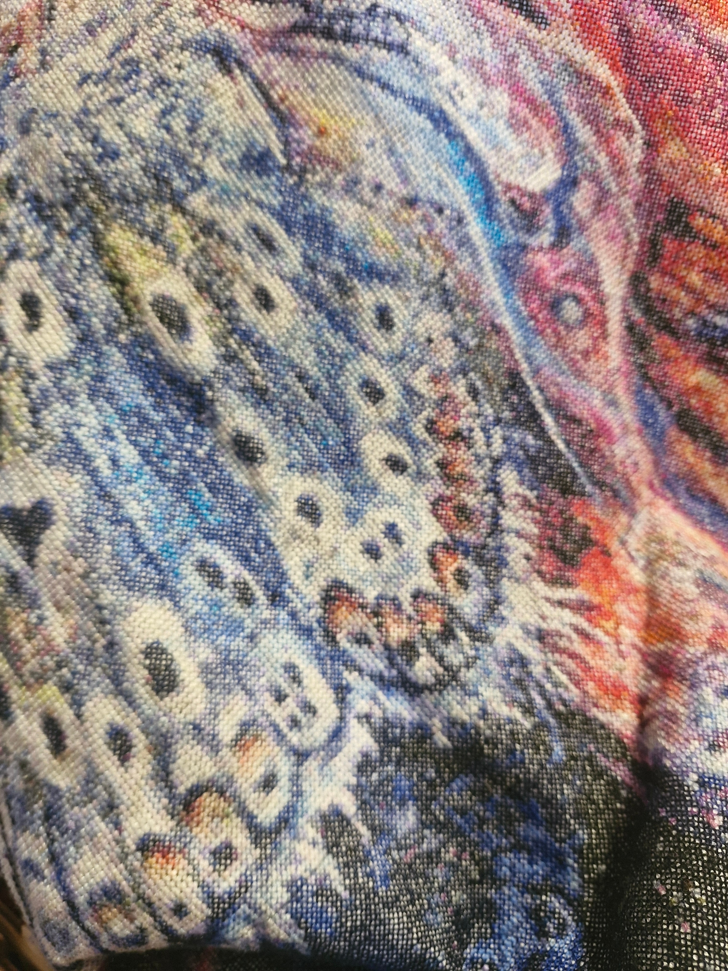 A close-up, angled view of a colorful, patterned textile is presented. The pattern features abstract shapes and swirling lines in shades of blue, red, pink, and yellow against a lighter background. The fabric appears to be woven, with visible threads creating a textured surface. The lighting is somewhat diffused, highlighting the intricate details of the print. The image captures a section of the fabric, with no discernible context of its location or surroundings.