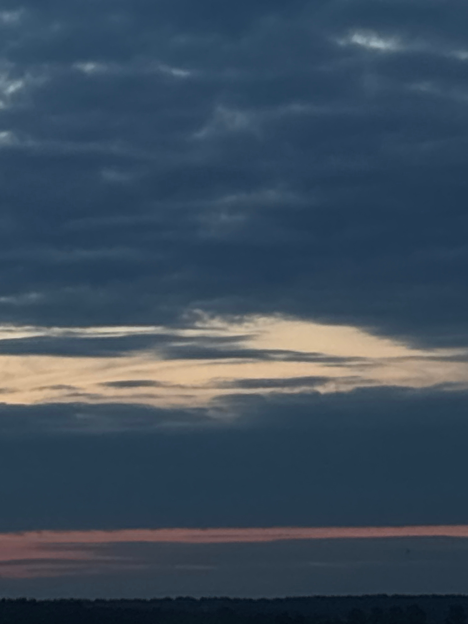 The image displays a vibrant sky scene, primarily composed of deep blue and grey tones, accentuated by dramatic cloud formations. The sky transitions from a darker blue-grey on the left to slightly lighter hues towards the center-right.
On the left side of the frame, there are scattered, diffuse patches of lighter grey and white clouds, suggesting a lower light intensity or thicker cloud cover in that area. These clouds offer subtle texture against the darker sky.
The most prominent feature is a broad, vertical band of brightly illuminated clouds traversing the central and right sections of the image. These clouds are elongated and streaky, glowing vividly in shades of white, cream, and soft golden yellow, indicative of direct sunlight catching them from a low angle. This strong illumination creates a stark contrast with the darker sky.
Further to the far right, a thin, distinct vertical streak of reddish-orange light is visible, likely another cloud formation or atmospheric effect catching the light differently. On the extreme right edge, a thick, dark vertical band, possibly a partial view of a structure or frame element, partially obscures the sky.
The dramatic lighting, with brightly illuminated clouds against a deep sky, strongly suggests either sunrise or sunset. The weather is partly cloudy. No people, specific objects (other than the potential frame element), or visible text are present in the sky. The location context is Minsk, Belarus, but no specific geographical cues are visible within the image itself.