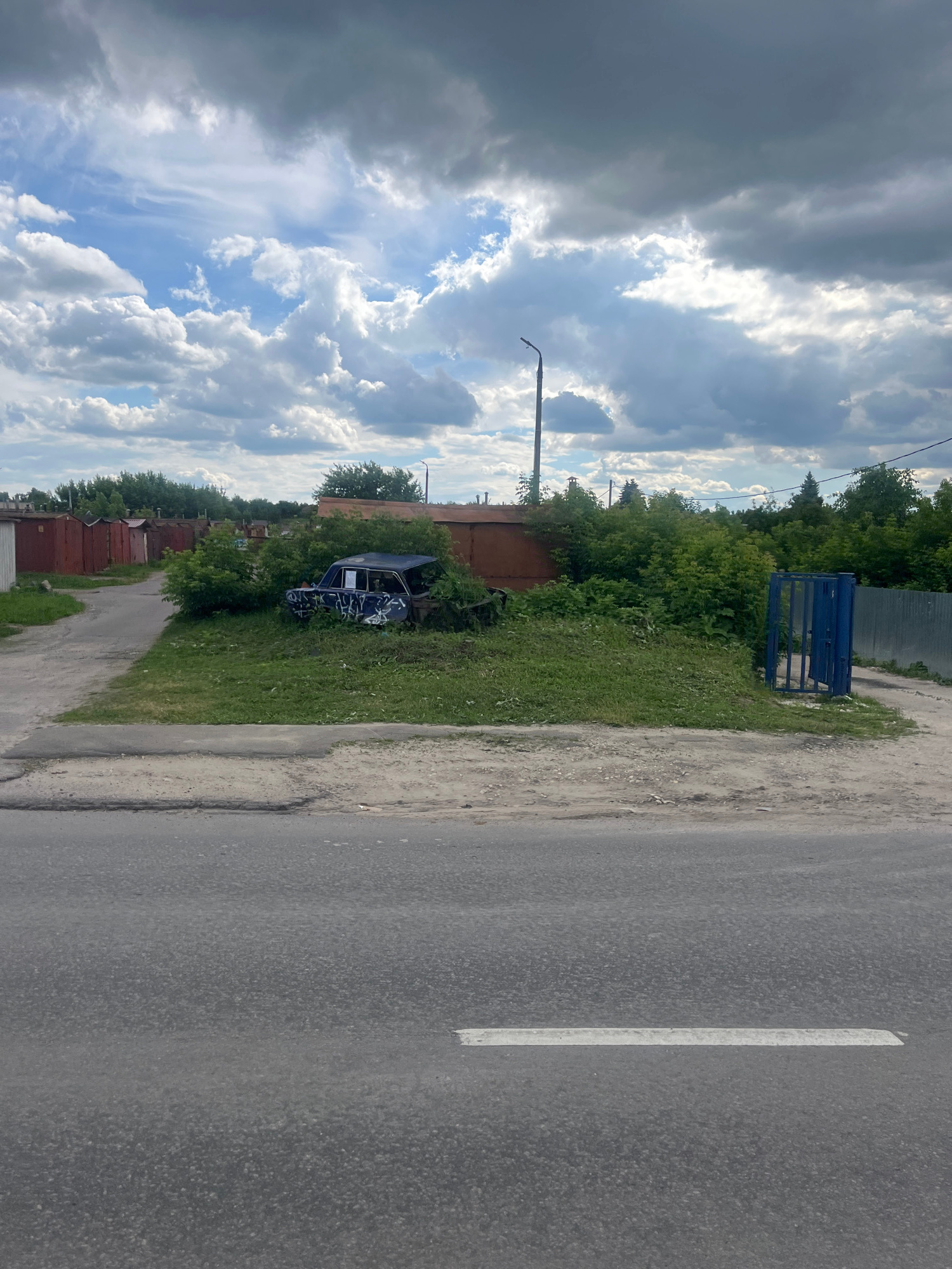 This is a vertically oriented, medium-shot photograph taken from a slightly elevated perspective. The scene depicts a street in Kolomna, Russia, likely during daylight given the bright sky. The sky is filled with dramatic, fluffy white and dark grey clouds, suggesting a dynamic weather pattern, possibly on the cusp of a storm or after rain.
The foreground is dominated by an asphalt road with a faint white line marking the lane. To the left of the road, there is a patch of overgrown grass and dirt, leading to a wooded area. Within this green expanse, a dark-colored car, possibly blue, is visible, partially obscured by foliage and covered in white graffiti. The car appears to be abandoned or wrecked, with its doors open.
Further left, behind the car, are several low-rise structures, possibly garages, with corrugated metal roofs. A rusty brown structure is prominent among them. Beyond the car and trees, a tall metal fence can be seen, and further in the distance, a blue metal gate or structure stands out against the greenery. The overall impression is one of an urban or semi-urban fringe area, perhaps an industrial or residential zone where older structures and natural overgrowth coexist. There is no visible text in the image. The scene conveys a sense of neglect and decay, particularly in the state of the car and the overgrown vegetation.