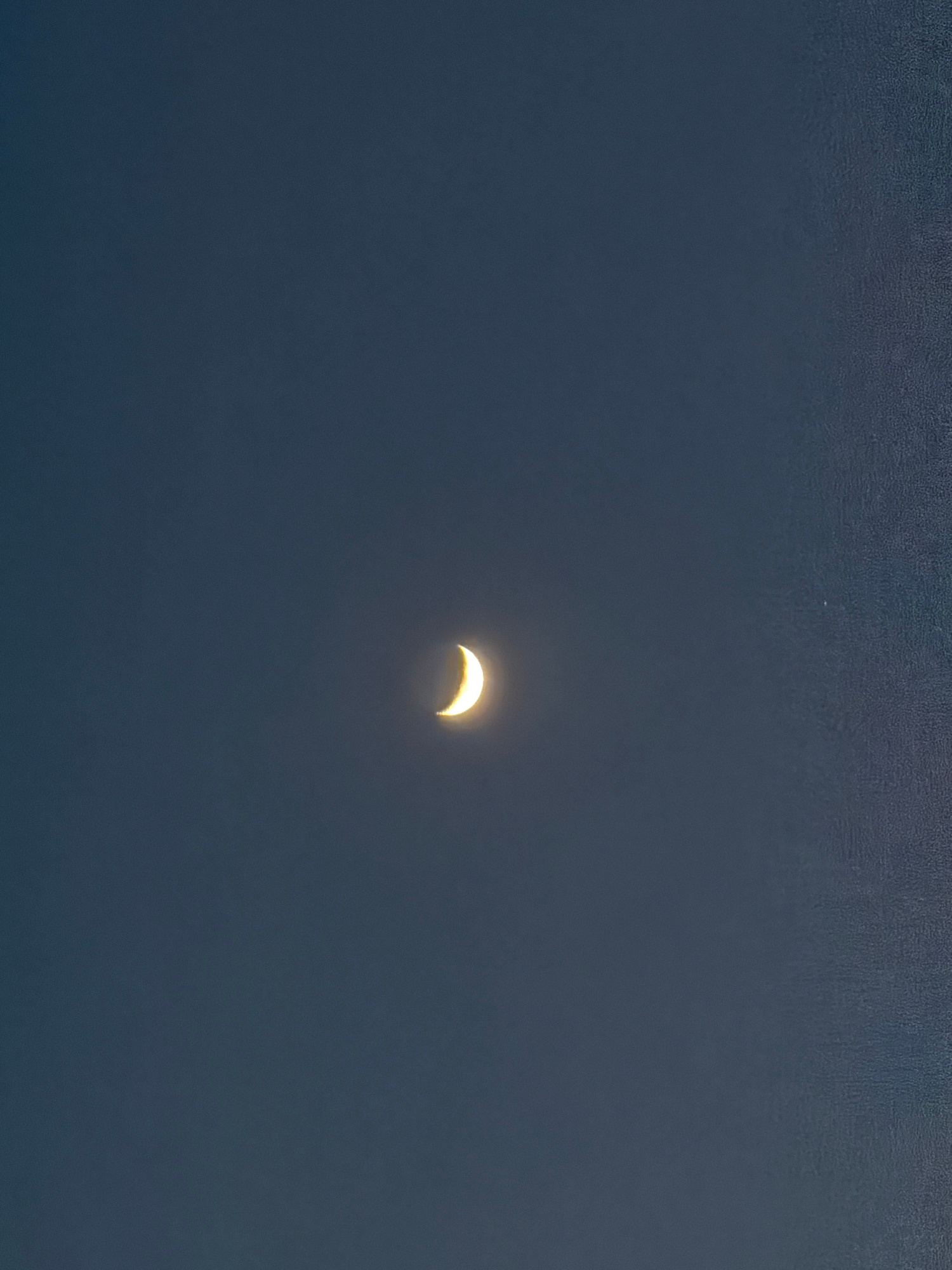 A crescent moon is visible in the dark sky. The moon is illuminated from the right and appears to be glowing with a warm yellow light. The sky is a deep, dark blue, with a slight texture suggesting a subtle grain in the image. The image is framed such that the moon is positioned slightly to the left of the center. There is a tiny white speck visible in the upper right quadrant of the image, possibly a star or a distant light source.