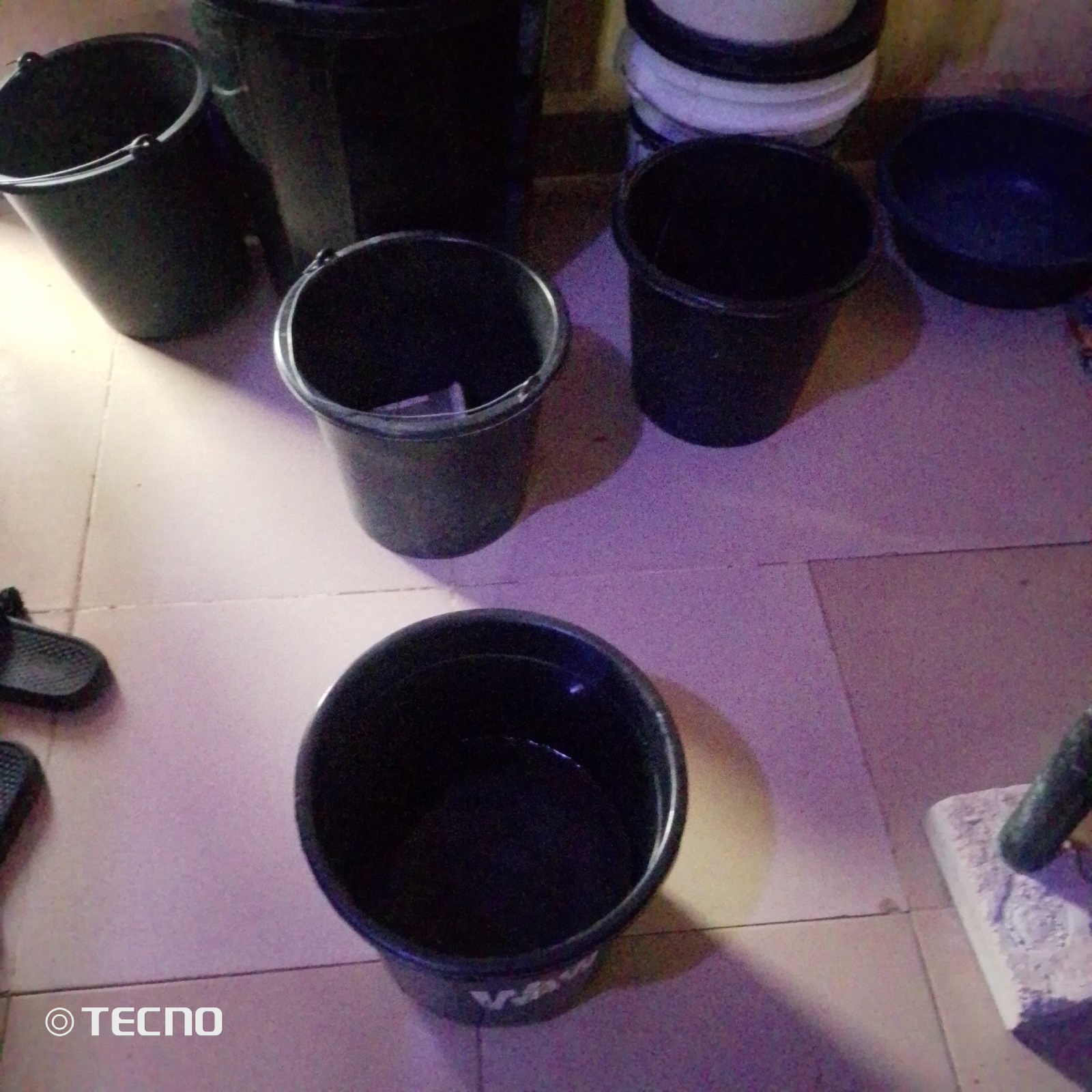The image depicts a tiled floor environment with several black plastic buckets and a blue basin scattered around. In the foreground, a large black bucket with some liquid inside is prominently displayed. To its right, a blue basin is partially visible. In the background, there are more black buckets, some stacked, and a white bucket is visible among them. A pair of dark-colored slippers are on the floor to the left of the image. The lighting suggests an indoor setting, possibly a storage area or a workshop. The image also contains a watermark "© TECNO" in the bottom left corner. The scene is located in Jalingo, Nigeria, a city known for its markets and local commerce, which could explain the presence of multiple buckets for various uses.