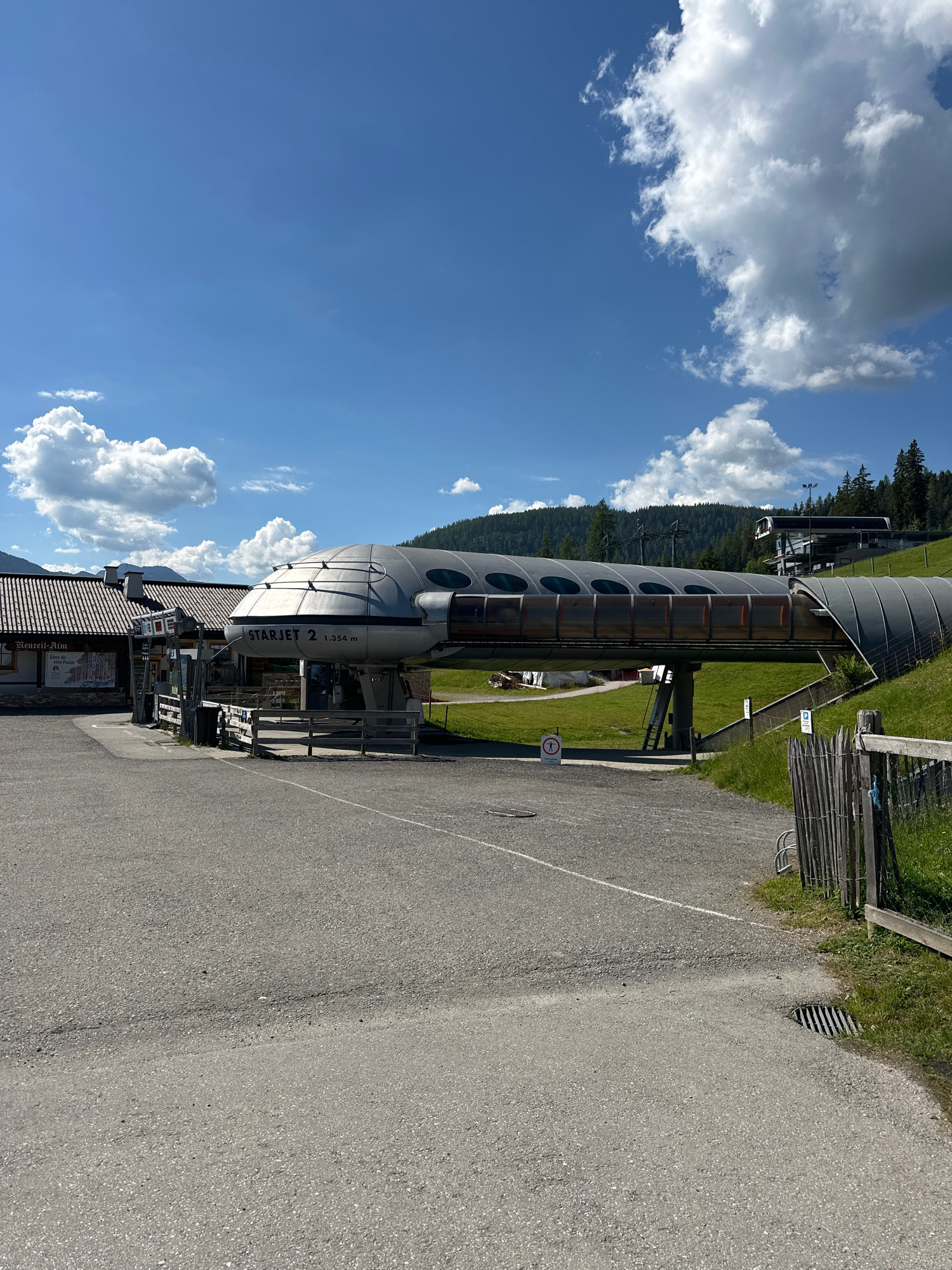 A ski lift station is visible under a bright blue sky with scattered white clouds. The ski lift has a futuristic, metallic design with a long, curved, covered walkway and a prominent "STARJET 2" label. To the left of the station, a building with a red tiled roof appears to be a restaurant or rest area, with signage visible. The area in front of the station is a paved lot, and to the right, a grassy slope with a wooden fence is present. The scene is set in a mountainous, likely alpine, region, with trees and hills in the background. The location is the STARJET 2 ski lift station in Flachau, Austria. There are no people visible in the image.
