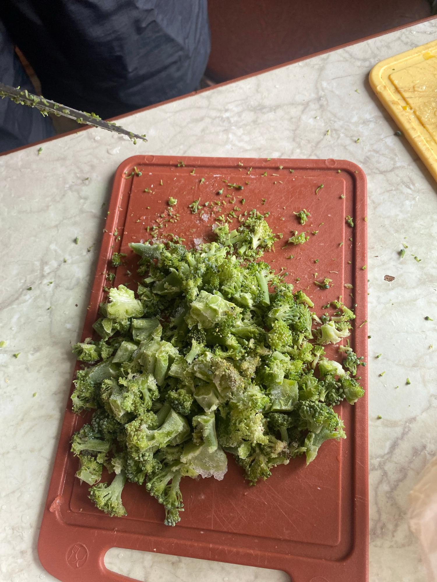 The image shows chopped broccoli on a red cutting board. The broccoli is fresh and green, with some small pieces scattered around the board. In the background, there is a person wearing a black jacket and a yellow cutting board. The scene appears to be a kitchen, possibly in a home. There is no information to determine the location in the city of Krupki, Belarus.