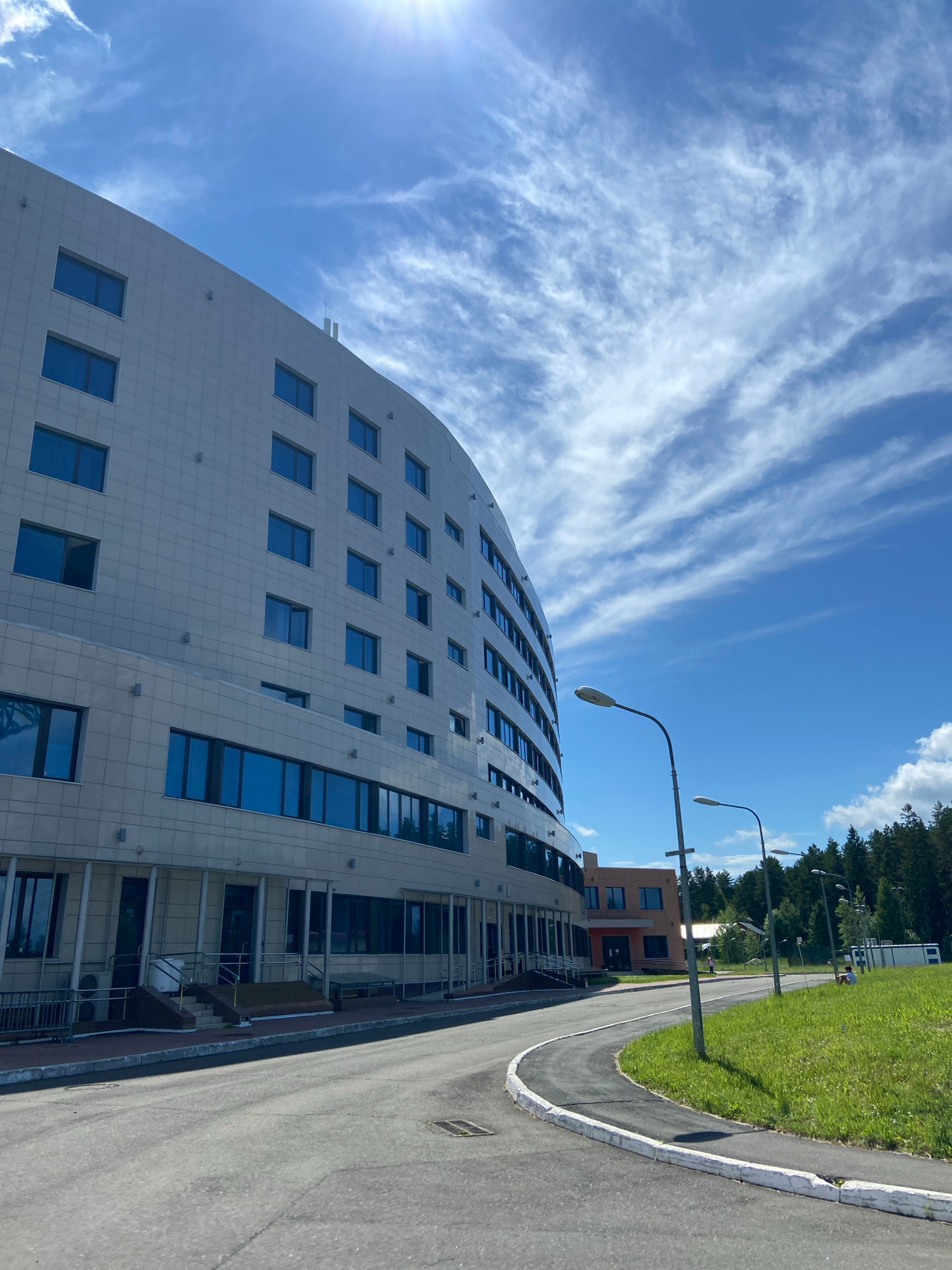 A modern, curved building with a tiled facade and numerous rectangular blue windows stands to the left of the frame. A paved road with a white curb curves from the foreground towards the building. Several lampposts line the road. Beyond the building and road, there is a green grassy slope leading up to a dense forest of pine trees under a bright blue sky with wispy white clouds and sun rays. In the distance, a smaller, orange building with dark windows is visible. A few people are scattered in the area, including one sitting on the grass and another walking near the orange building.