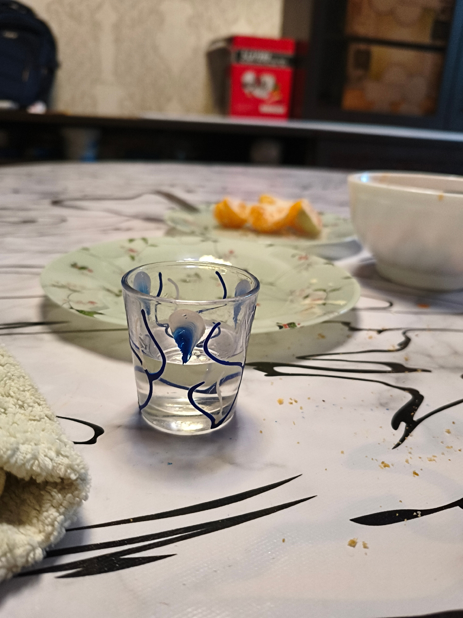 The image shows a close-up of a table surface with various objects. In the foreground, a small glass containing a clear liquid and decorated with blue swirls is prominent. To the left of the glass, a white fluffy cloth is visible. Behind the glass, a plate with what appears to be slices of orange is partially visible. Further in the background, a white bowl and a blurred red object are present. The table surface has a white, marble-like pattern with black abstract designs and scattered crumbs. The lighting suggests an indoor setting, possibly a dining area. There is no indication of the city of Rudny, Kazakhstan.