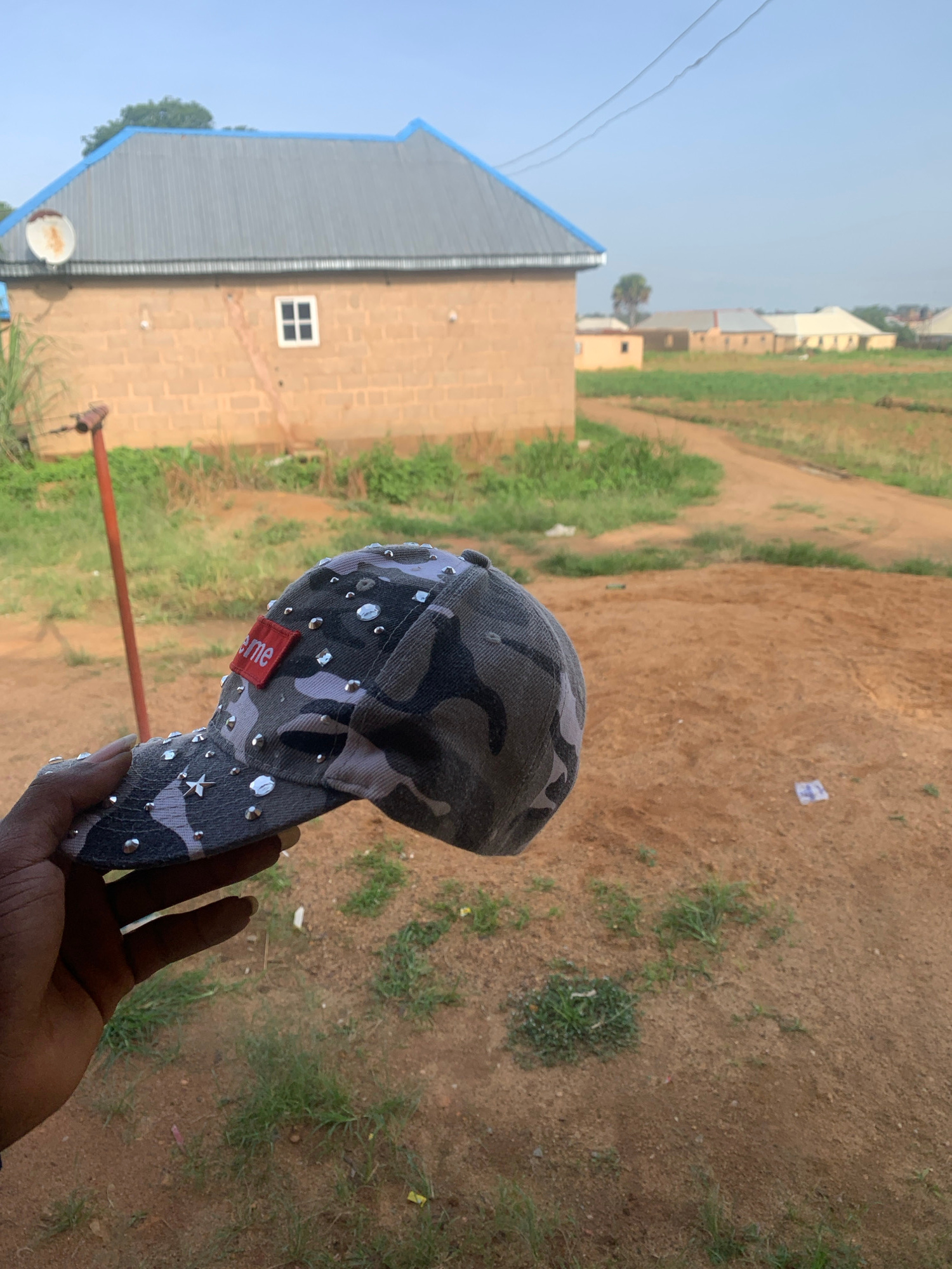 A person's hand is holding a camouflage-patterned baseball cap adorned with rhinestones and a red rectangular tag with white lettering. The cap is held in the foreground, angled towards the right. In the background, there is a light brown brick house with a corrugated metal roof. A single square white-paned window is visible on the side of the house. The environment appears to be a rural or semi-rural area in Bukuru, Nigeria, with dirt paths and sparse vegetation. In the distance, other buildings with similar roofs are scattered amongst fields under a clear sky. The overall scene suggests an outdoor setting, possibly a residential area.