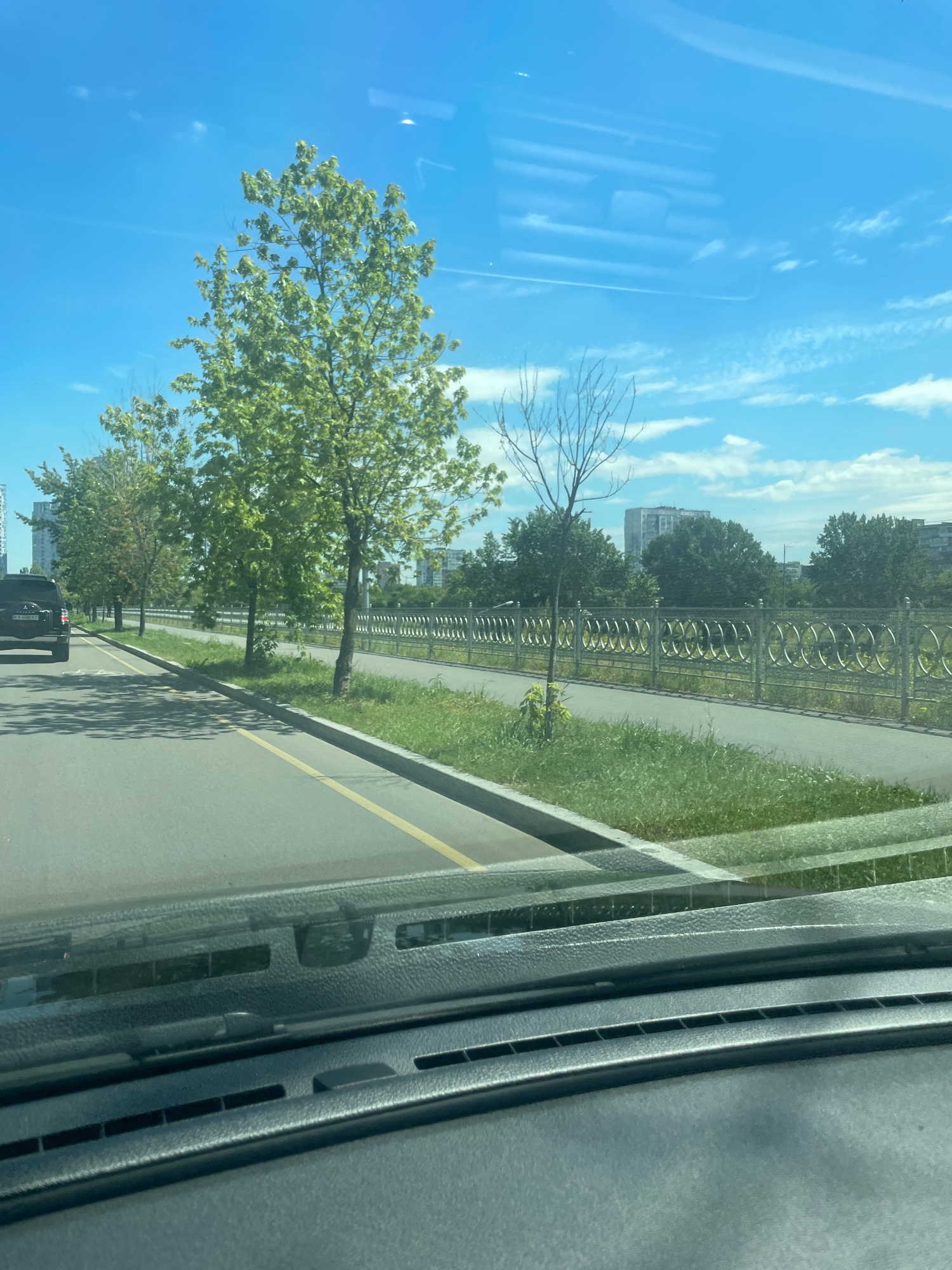 The image shows a view from a car interior, looking out through the windshield at a street scene in Kyiv, Ukraine. A black SUV is driving on the left side of the road, which has a yellow lane marker. Trees line the road on the right, with a paved walkway behind them, bordered by a decorative metal fence. In the background, tall buildings and more trees are visible under a bright blue sky with scattered clouds. The foreground of the image shows the dashboard of the car, including the windshield wiper mechanism and air vents.