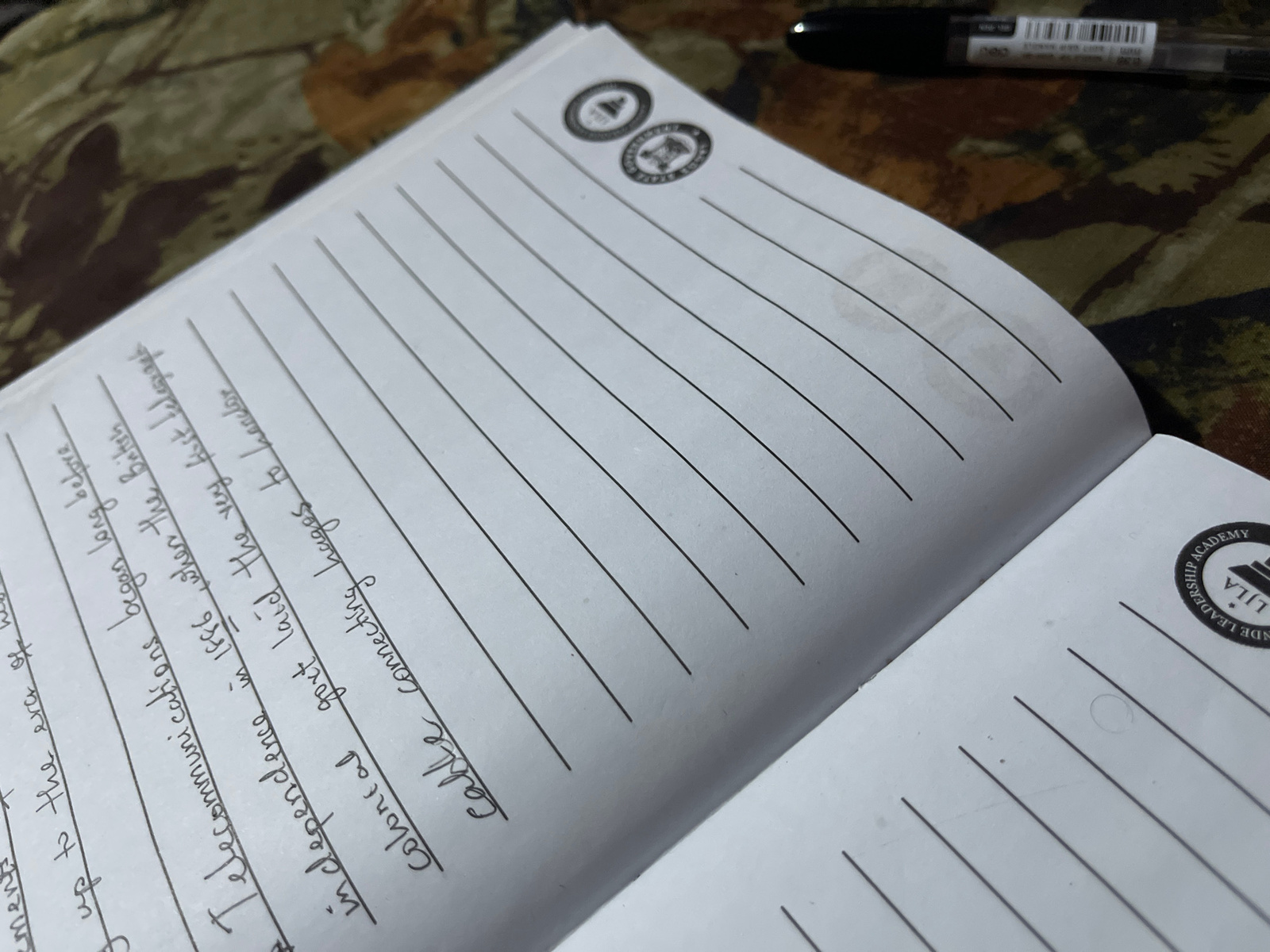 The image shows an open notebook with lined pages. There are two circular emblems at the top of the left page, each with a design inside. Handwritten text is visible on the left page, starting with "The British" and continuing with "The telecommunications" and "in dependence" followed by "cable". A black pen is positioned horizontally in the upper right corner of the image, resting on a patterned surface that appears to be fabric, possibly a camouflage-patterned cloth. On the right page, a circular emblem with the words "LILA LEADERSHIP ACADEMY" is visible. The notebook is placed on the patterned fabric surface. There is no indication of the city of Alasia, Nigeria, or any specific urban environment.
