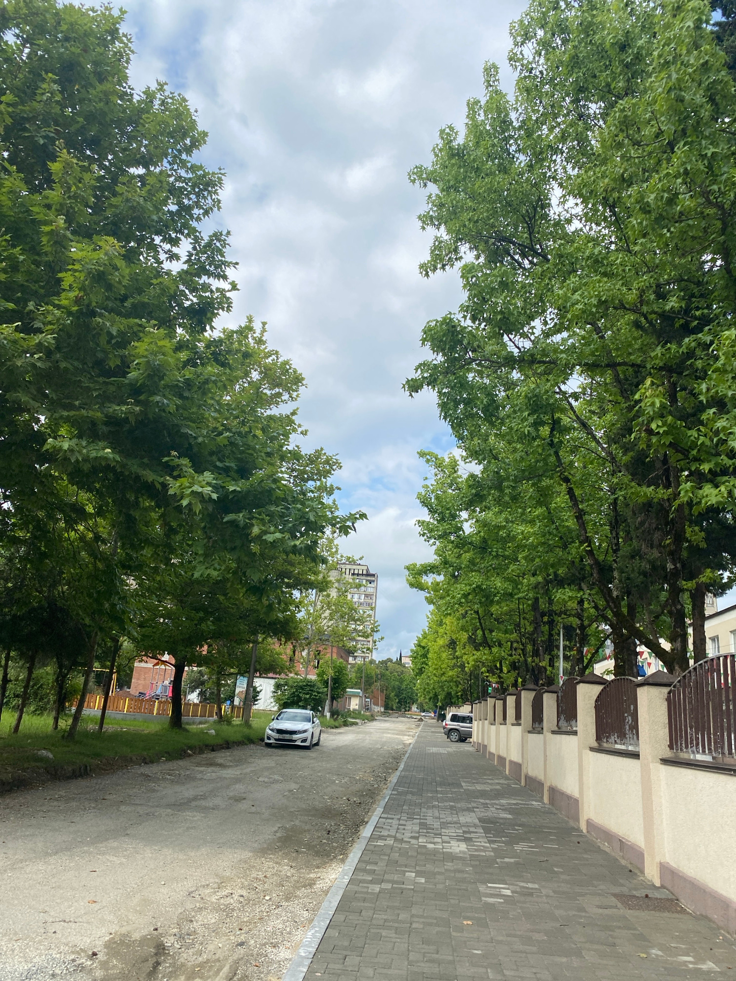 A street scene unfolds under a partly cloudy sky. Tall trees with lush green foliage line both sides of a paved sidewalk, creating a shaded canopy. A dirt and gravel road runs parallel to the sidewalk, with a white car parked on the left side. Further down the road, another car is visible. A decorative fence with brown posts and a cream-colored wall runs along the right side of the sidewalk. In the background, buildings of varying heights, including a tall, multi-story structure, are visible. A playground with yellow fencing and red equipment is situated on the left side of the road. The overall impression is a quiet, tree-lined street in a residential or urban area.