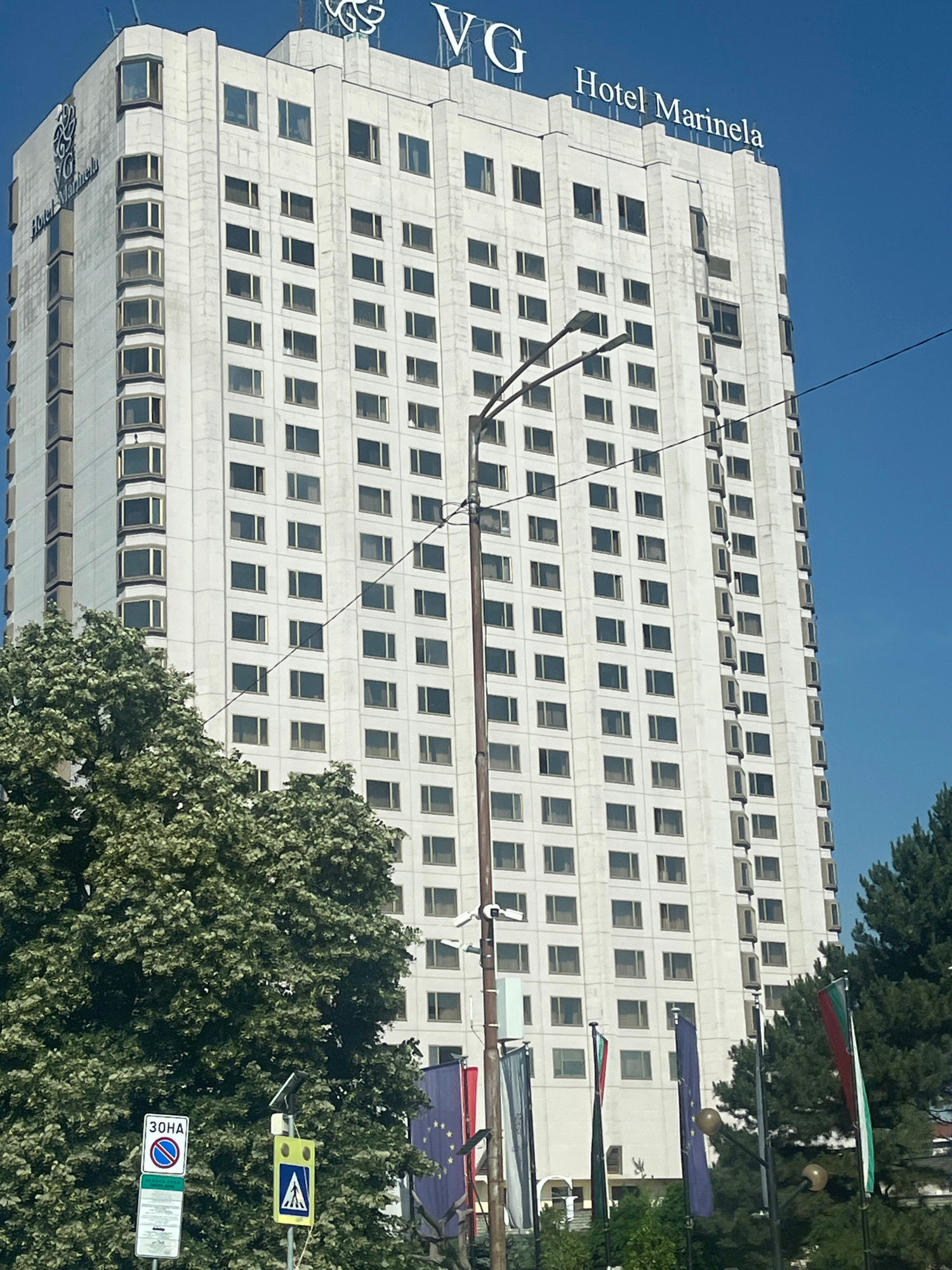 A tall, white, rectangular skyscraper dominates the frame, featuring a grid of numerous windows. The top of the building displays the text "VG Hotel Marinela". In the foreground, lush green trees partially obscure the lower portion of the building and the street. Several flags are visible, including what appears to be the European Union flag and other national flags. Traffic signs are also present, including one with Cyrillic text reading "30HA". A lamppost with multiple lights is positioned in front of the building, and a power line extends diagonally across the scene. The sky is a clear, bright blue.
