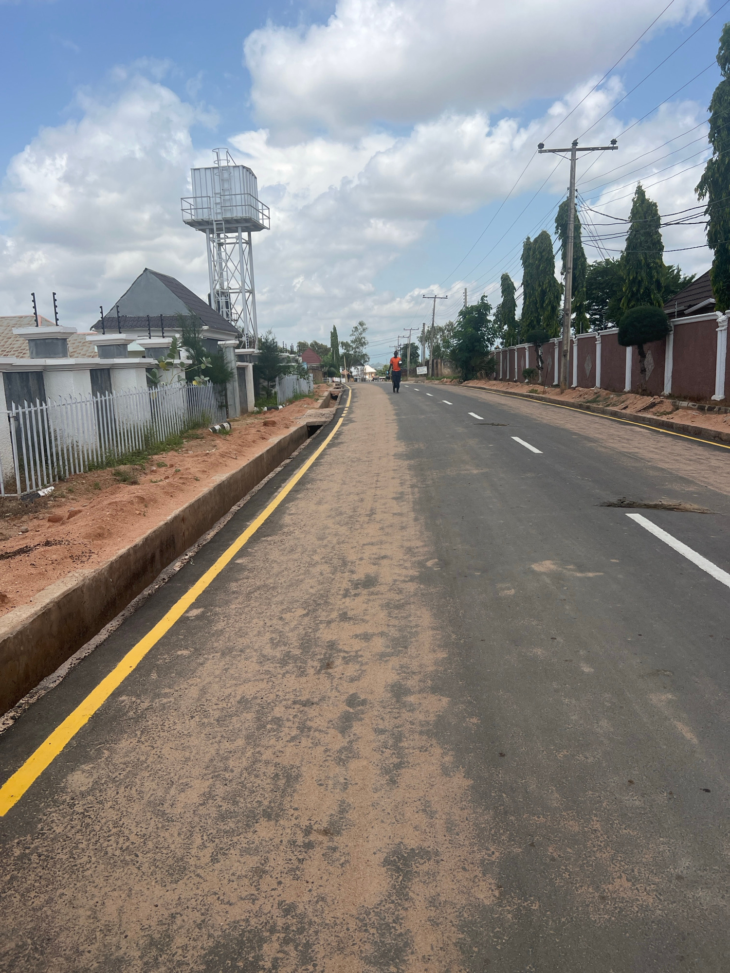 The image depicts a street scene in the city of Howayi, Nigeria. A person is seen jogging down the center of the road, which is paved and has white lane markings. To the left of the road, there is a drainage ditch with a yellow line painted along its edge. Beyond the ditch is a white fence with decorative pillars, and behind that, a house with a prominent water tower. Tall, slender trees line the right side of the road, separating it from more residential structures. The sky is partly cloudy.