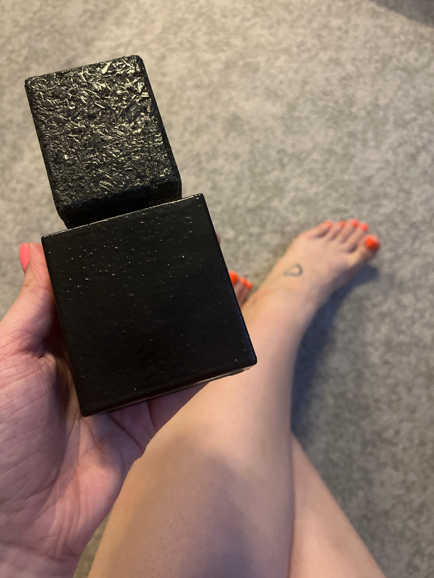 An overhead, high-angle shot shows a person's hand holding a black, rectangular object with a textured lid that is slightly ajar. The object appears to be a perfume bottle. To the right of the bottle, a person's bare foot is visible with bright orange toenail polish and a small, faint tattoo on the ankle. The background is a textured gray carpet. There are no identifiable landmarks or features to suggest a specific location within Riga, Latvia.