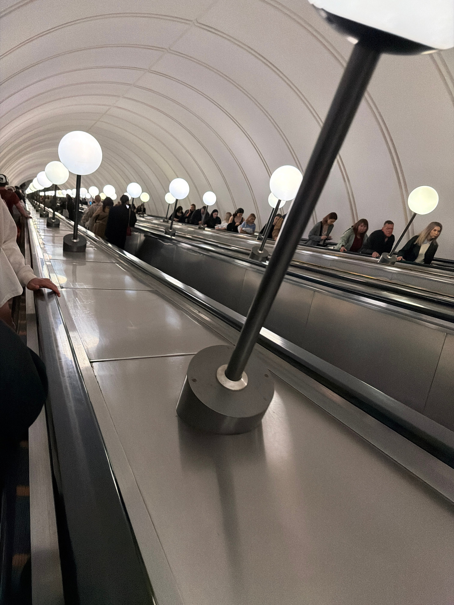 This is a photograph taken from a downward-facing perspective, looking along an escalator in what appears to be a subway station. A row of globe-shaped lights illuminates the curved ceiling of the tunnel, creating a tunnel effect. Several people are visible on the escalator, ascending or descending, their faces indistinct due to the angle and distance. A dark railing runs along the side of the escalator, with metallic components visible at intervals. The overall impression is of a busy, functional urban transit space. The lighting suggests it could be any time of day, and there are no clear indicators of weather conditions. The scene is devoid of any visible text.