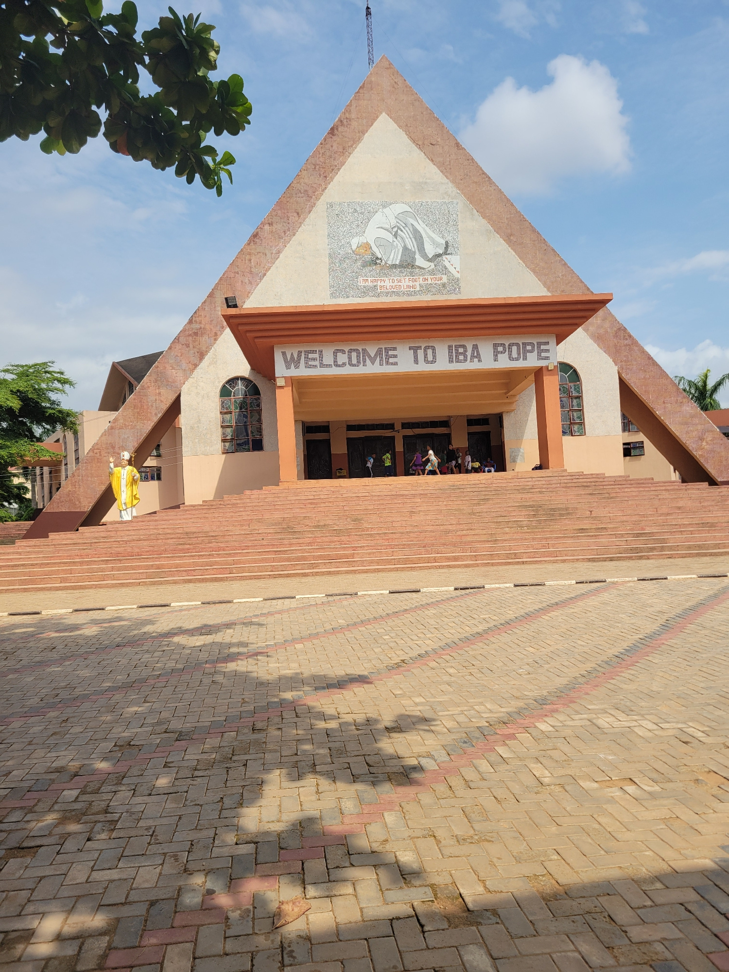 A full shot shows the front of a church with a large, peaked roof. The roof is adorned with a mosaic of a kneeling figure and text that reads "I AM HAPPY TO SET FOOT ON YOUR BELOVED LAND." Below the mosaic, a sign reads "WELCOME TO IBA POPE." A wide set of stairs leads up to the entrance. A statue of a figure in a yellow robe and white mitre stands on the left side of the stairs. Several people are gathered near the entrance. The foreground is a paved area with a pattern of interlocking bricks. Trees and a blue sky with clouds are visible in the background.