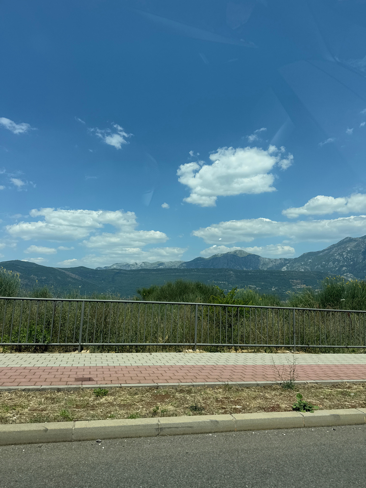 The image depicts a scenic view of a mountainous landscape under a bright blue sky with scattered white clouds. In the foreground, there is a paved sidewalk with a brick pattern, bordered by a metal fence and a roadside verge with dry grass and a small green plant. Beyond the fence, lush green vegetation covers the lower slopes of the mountains. The mountains themselves are rugged and rocky, with some trees and sparse vegetation visible on their slopes. The sky is clear and blue, suggesting a sunny day. There are reflections in what appears to be a window, possibly indicating the photo was taken from inside a vehicle or building. The overall scene evokes a sense of peace and natural beauty. The time of day appears to be daytime, likely late morning or afternoon given the bright sunlight. The location is identified as Đuraševići, Montenegro. There is no visible text in the image.