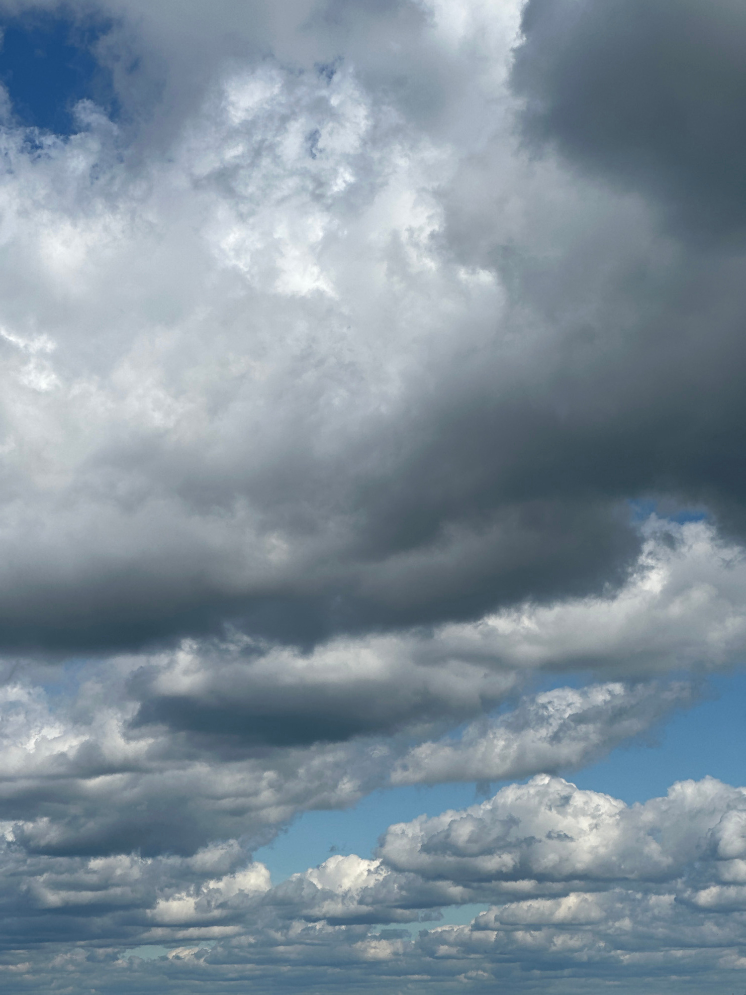 A vertical, medium shot captures a sky filled with a dramatic and varied cloudscape. The image is dominated by shades of gray and white, with only small patches of bright blue sky visible.
In the upper portion of the image, a deep blue opening in the clouds reveals a clear sky. This blue expanse is surrounded by a dense, white, and grey cloud formation that stretches across the top half of the frame. The clouds here appear fluffy and textured, with some areas catching the light and appearing almost luminous, while others are darker and more shadowed, suggesting depth and density.
Moving down, the cloud cover becomes thicker and darker. Large, ominous-looking grey clouds occupy the mid-section of the image, obscuring much of the sky. These clouds have a more uniform and heavy appearance, with subtle variations in tone indicating their mass.
Towards the bottom of the frame, the sky opens up again, revealing patches of brighter blue. Here, a layer of white, puffy clouds with well-defined edges are visible, contrasting with the darker clouds above. These lower clouds are illuminated from above, creating a sense of dimension and layering. At the very bottom of the image, a faint, hazy horizontal line suggests a distant horizon, possibly a cityscape or landscape, but it is too indistinct to be clearly identified.
The overall impression is one of a dynamic and changing sky, possibly indicative of impending weather or the aftermath of a storm. The lighting suggests it is daytime, but the heavy cloud cover creates a diffused and somewhat moody atmosphere. There are no people, animals, or man-made objects clearly depicted within the frame, focusing solely on the natural element of the sky and its cloud formations. The location is unknown.