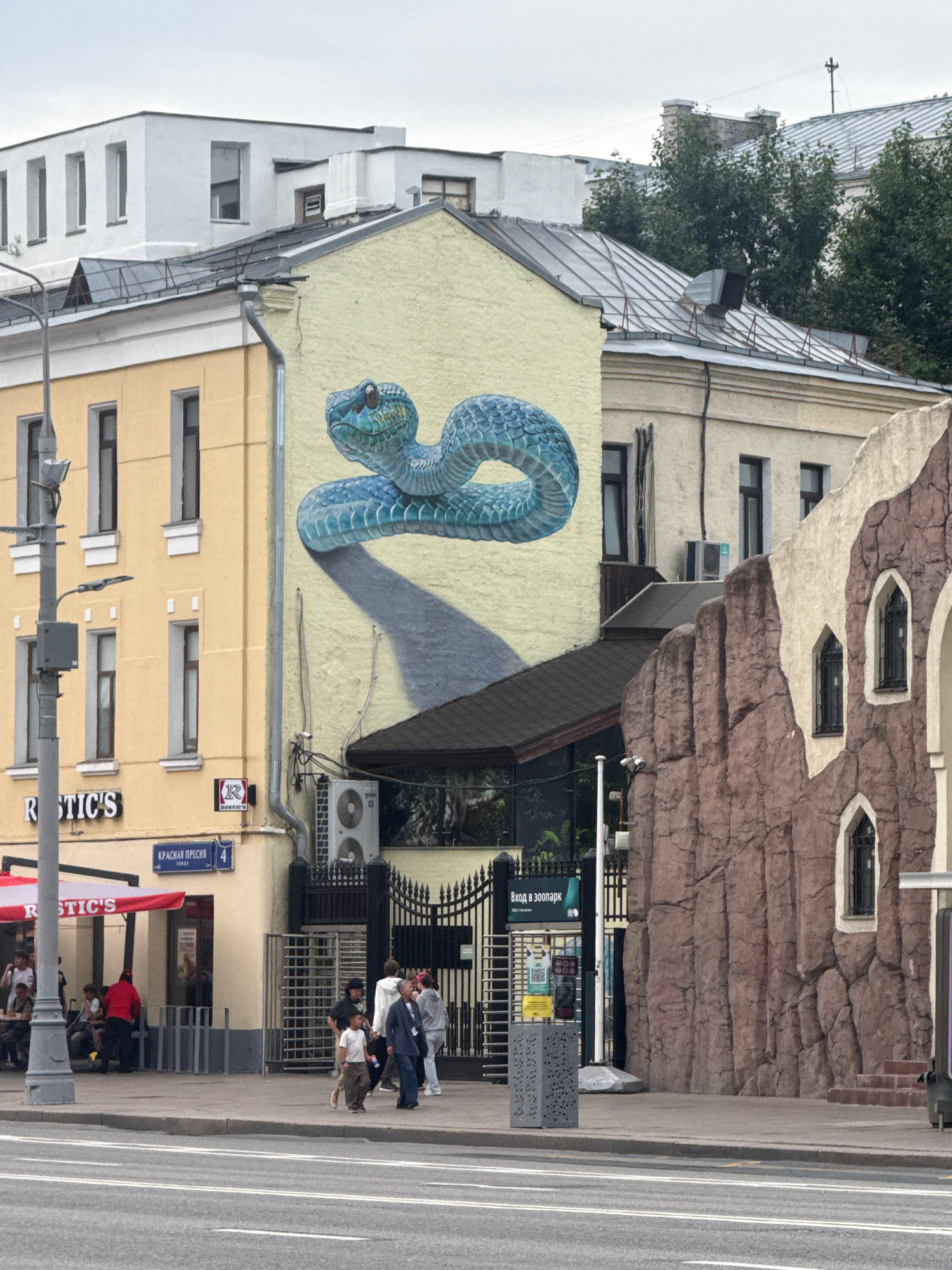 The image depicts a street scene in Moscow, Russia, featuring a building with a large mural of a blue snake on its facade. The building on the left is yellow with several windows and a sign that reads "RUSTIC'S". A red awning extends over an outdoor seating area. Further to the right, a building with a textured, rock-like facade is visible, with arched windows. Several people are seen walking on the sidewalk and gathered near the entrance of what appears to be a zoo or park, indicated by signs with Russian text that translate to "Entrance to the Zoo". The weather appears overcast.