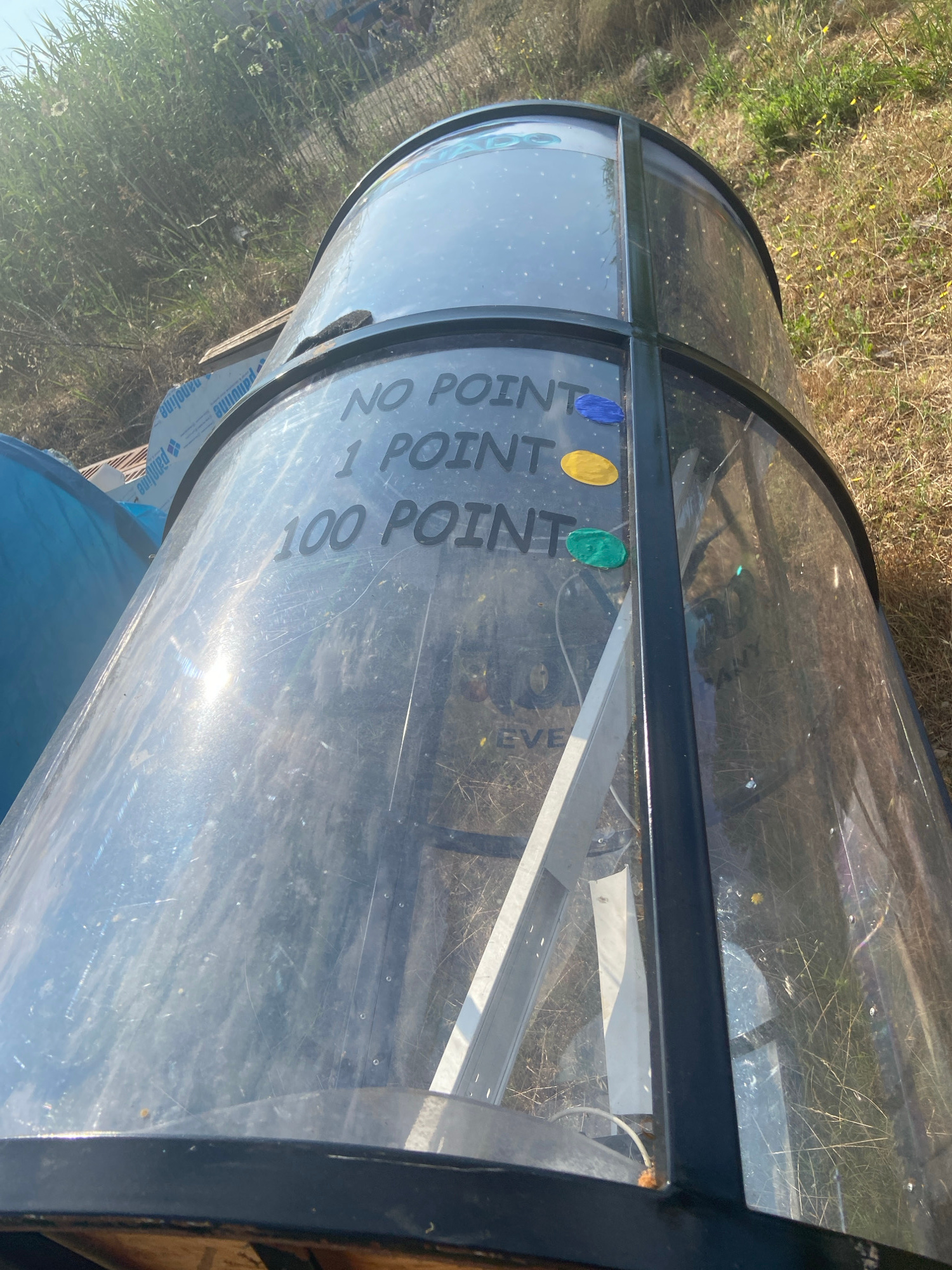 A transparent cylindrical object with black metal framing, possibly a game or prize dispenser, is located outdoors on dry, grassy terrain. The object has text printed on its surface reading "NO POINT", "1 POINT", and "100 POINT". Three colored circular decals, blue, yellow, and green, are affixed near the text. The scene is illuminated by bright sunlight, casting reflections on the object. In the background, tall green reeds and dry grass are visible, suggesting a natural or semi-rural environment. No people are present in the frame.