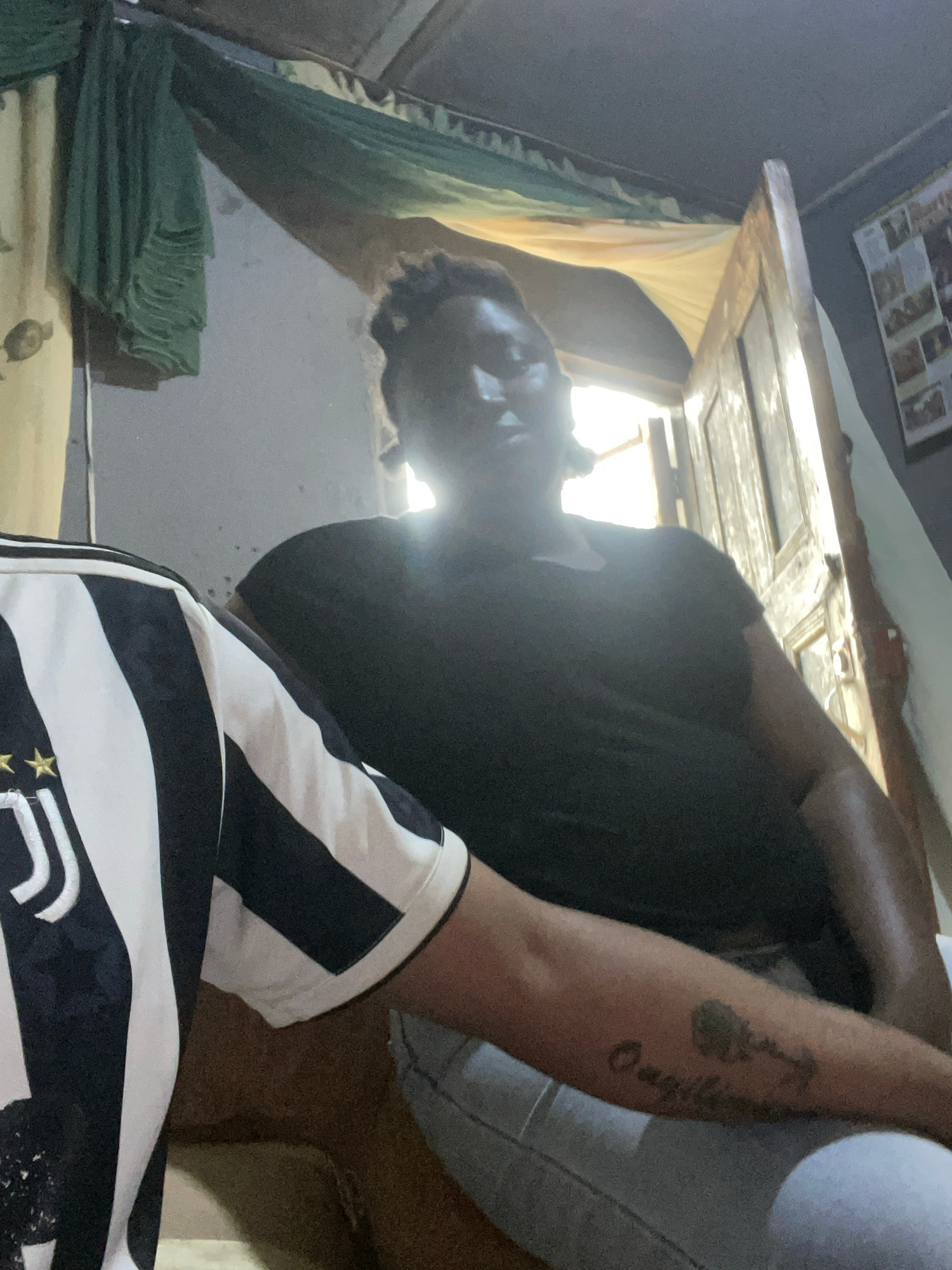 In Jos, Nigeria, a person wearing a black and white striped football jersey is captured in a selfie. The background suggests an indoor setting with a door and hanging fabric. The person's arm, visible in the foreground, displays a tattoo. The lighting in the image is dim, with a bright light source emanating from behind the subject, creating a silhouette effect on their face. The overall ambiance is casual and personal, indicative of a domestic environment.