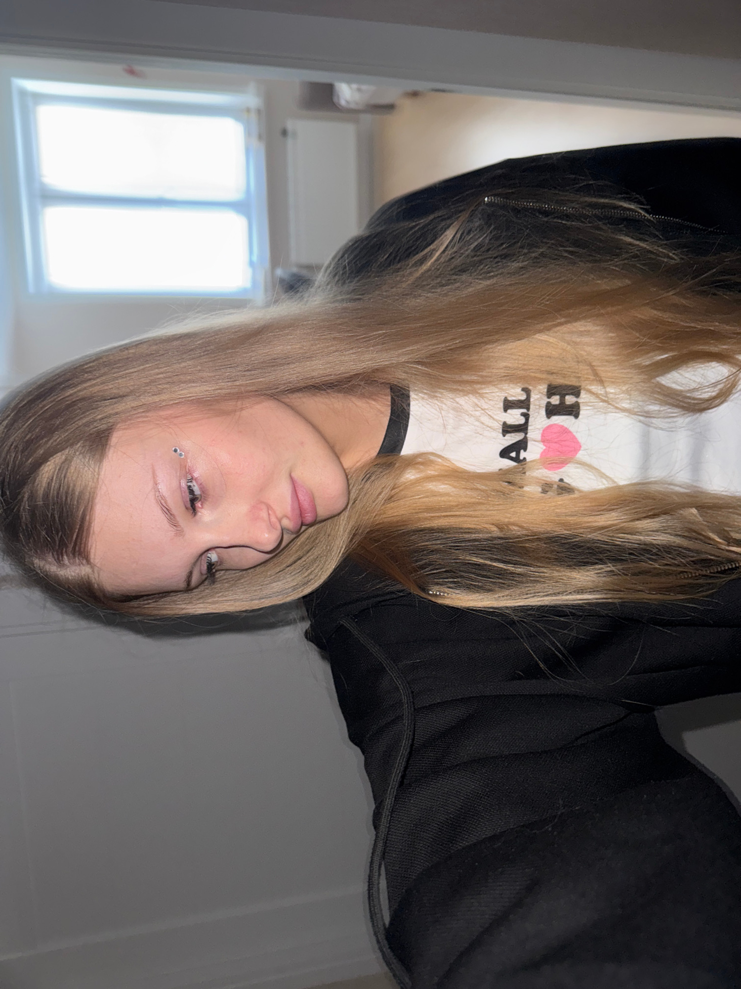 The image shows a young woman with long blonde hair and a black jacket. She is wearing a white t-shirt with black trim and text that appears to say "ALL I H". She has two small, clear rhinestones applied to her left eyebrow. The background is out of focus but appears to be an indoor setting with a window on the left side of the frame. The image is oriented sideways, with the woman's head on the left side of the frame. There is no discernible information about the city of Perm, Russia, or any specific urban location.