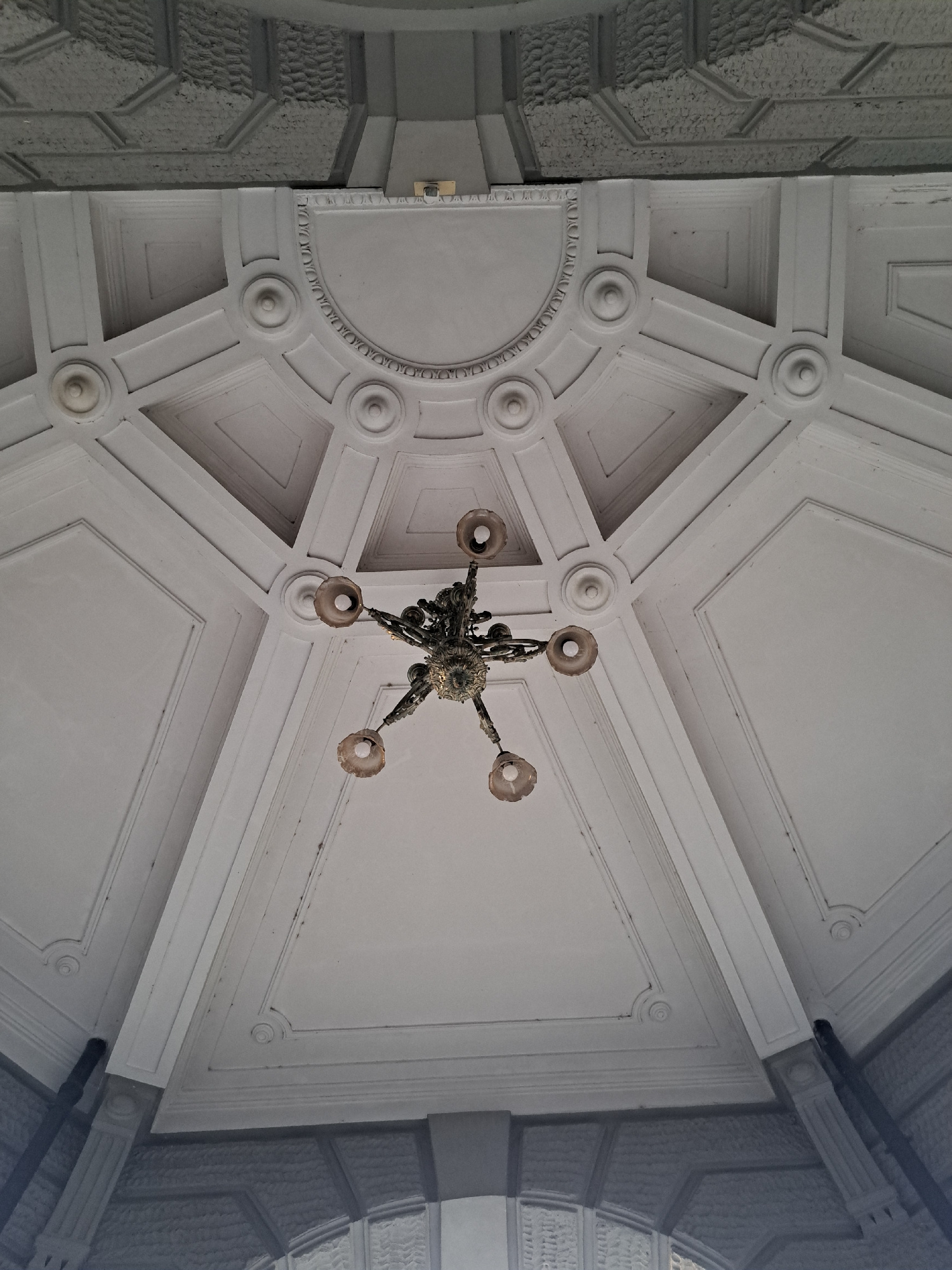 The image is a low-angle shot looking up at an ornate, white, coffered ceiling with a vintage chandelier hanging in the center. The ceiling is intricately decorated with geometric patterns, circular motifs, and raised molding. The chandelier is dark-colored and has five arms, each ending in a frosted glass shade, though the bulbs inside appear to be lit. The overall impression is one of classical architecture and grandeur. The scene appears to be indoors, likely in a public building or historical site. There are no people or any other visible elements in the image.