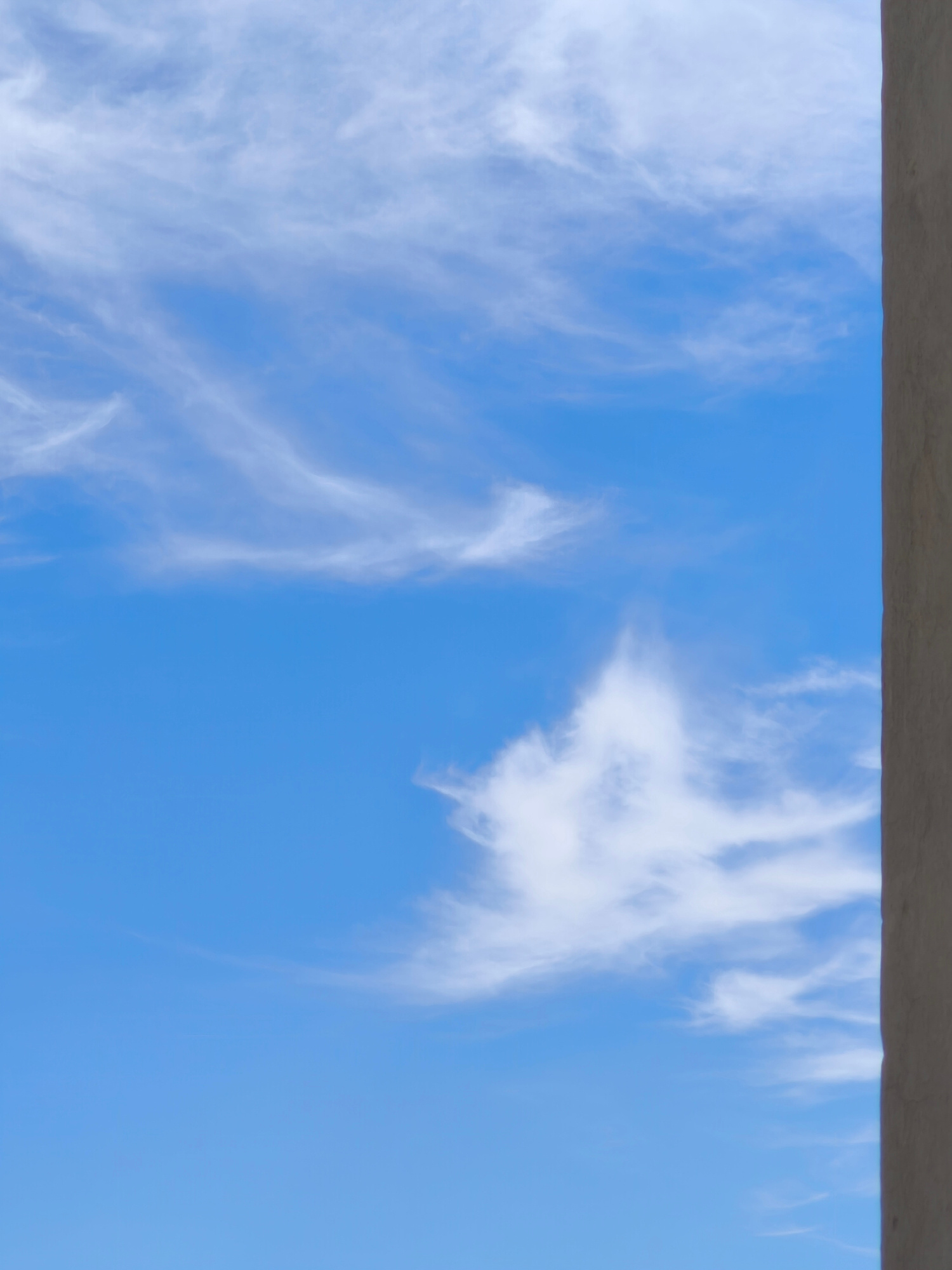 This is a vertically oriented, eye-level, medium shot of a bright blue sky filled with thin, wispy white clouds. The clouds are scattered and appear to be high in the atmosphere, suggesting fair weather. To the right of the frame, a textured, light brown or beige surface occupies a significant portion of the edge, appearing to be a wall or architectural element. The lighting indicates it is daytime, likely with bright sunlight illuminating the sky and clouds. There is no visible text in the image. The scene evokes a sense of openness and tranquility, with the vast sky dominating the composition. The wall on the right provides a grounding element and a sense of perspective, framing the expansive sky. The lack of any discernable human presence or activity suggests a peaceful and still moment.