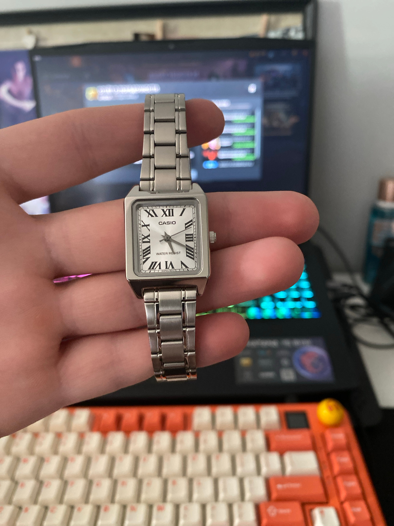 A person's hand is holding a silver-toned Casio watch with a rectangular face and Roman numerals against a blurred background. The background shows a laptop screen displaying what appears to be a video game or application, and a keyboard with orange and white keys. There is no information in the image to indicate the location in the city of Łódź, Poland.