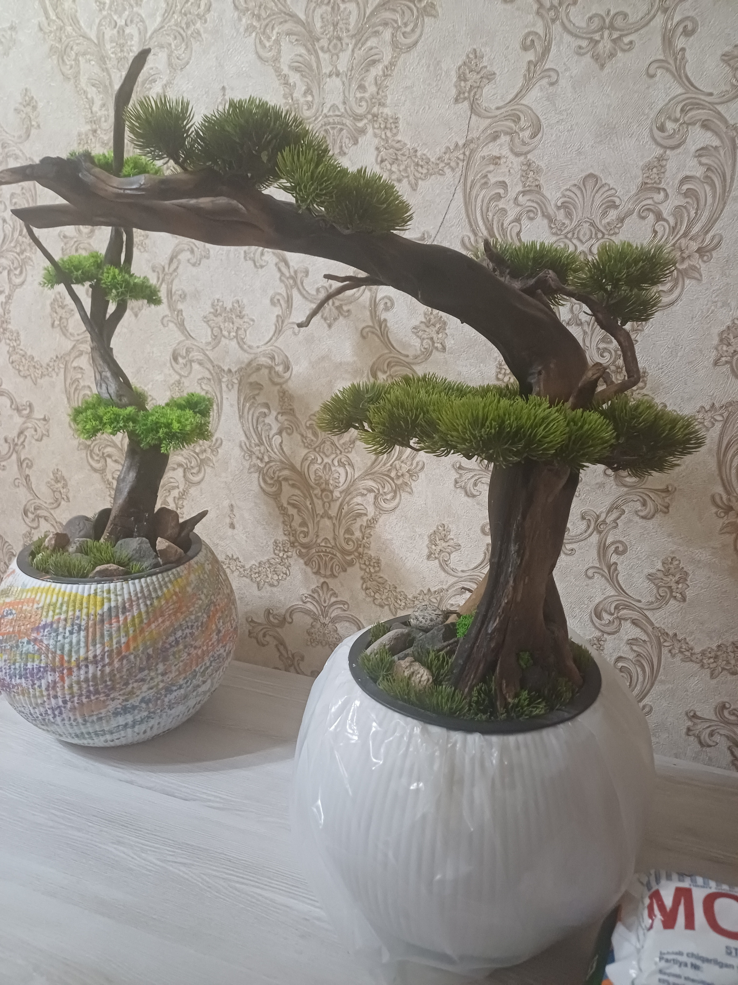 The image shows two artificial bonsai trees in decorative pots placed on a white wooden surface. The background is wallpaper with an ornate damask pattern in light beige and cream tones.
The bonsai trees appear to be made of plastic and are designed to mimic coniferous trees, with dense, green foliage clustered on branches. The trunks and branches are a dark brown, resembling aged wood.
The pot on the left is spherical and has a textured surface with a multicolored, almost mosaic-like pattern in shades of orange, yellow, blue, purple, and white. This pot contains a smaller bonsai tree, with small rocks and green artificial moss or grass at its base.
The pot on the right is also spherical, but it is covered in white plastic wrapping, giving it a smooth, slightly wrinkled appearance. This pot holds a larger bonsai tree with a more prominent trunk and sprawling branches. The base of this bonsai is also decorated with small rocks and green artificial grass.
In the lower right corner of the image, a portion of a white plastic package with red lettering is visible. The visible letters are "MO" and possibly part of another word.
The lighting in the image is soft, creating subtle shadows on the surface. The scene appears to be an indoor setting, possibly a home or a shop display, given the decorative wallpaper and the placement of the objects. No people are visible in the image. There is no information within the image to suggest a specific location in Tashkent, Uzbekistan.