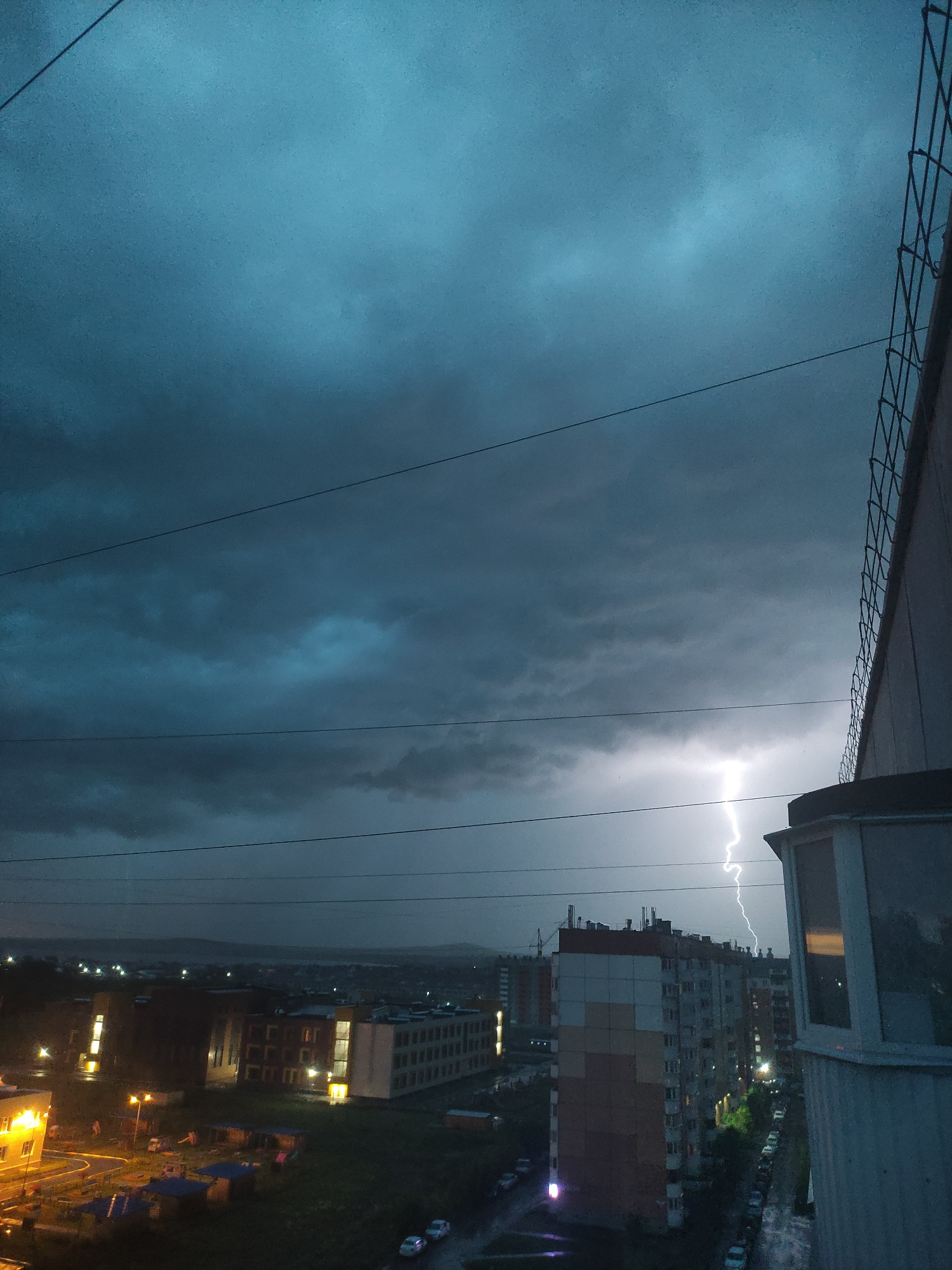 An image taken from an elevated perspective, likely a balcony or window, shows a city skyline at dusk or during a storm. The sky is a turbulent, dark blue with heavy clouds. A prominent lightning strike illuminates a portion of the sky in the mid-ground. In the foreground, the edge of a building with a wire fence is visible on the right. Below, city buildings, some with illuminated windows, and a street with parked cars are visible. The scene is set in Magnitogorsk, Russia, as indicated by the context provided. There are no discernible people or specific actions depicted. The environment appears to be urban and experiencing stormy weather.