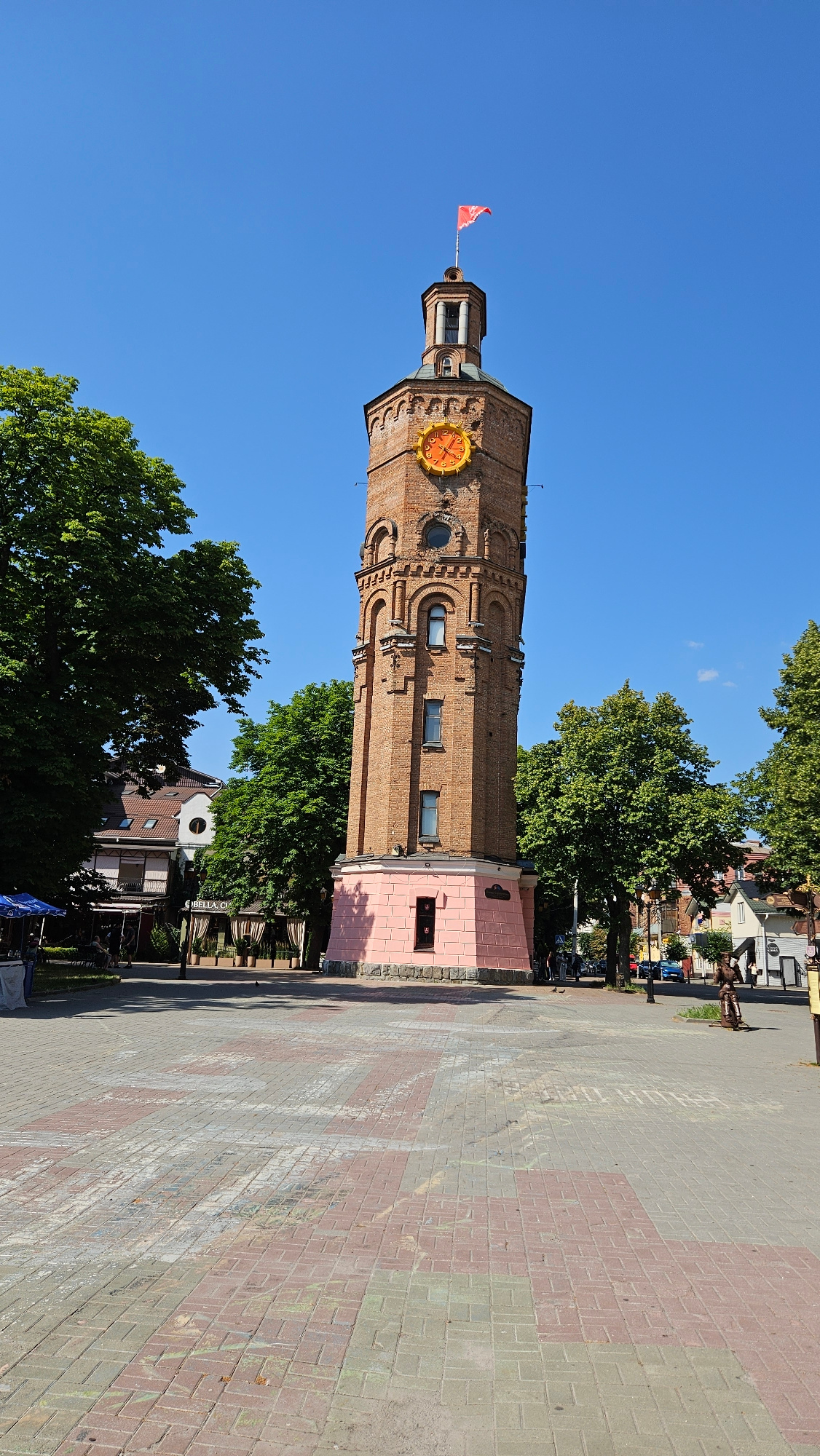 The image shows a tall, brick clock tower on a sunny day. The tower has an orange clock face with red hands. A small red flag flies from the top of the tower. Next to the tower is a small, pink building with a dark window. The ground in front of the tower is paved with red and gray bricks. Trees are visible around the tower, and in the background, there are some buildings and a street with cars. The sky is clear and blue. The image is oriented vertically, making it appear as if the tower is lying on its side.
