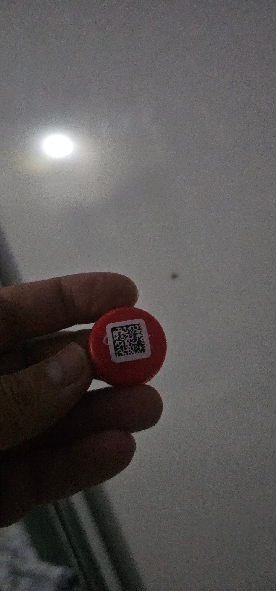 A hand holds a red circular object with a QR code sticker on it. The object is positioned in the center of the frame, and the hand is gripping it from the sides, with the fingers of the thumb and index finger visible. The background is a light grey, textured surface, possibly a ceiling, with a bright, out-of-focus light source in the lower left quadrant, suggesting artificial lighting. A small, dark speck is visible in the upper center of the background, which could be a dust particle. The overall lighting is dim, with a strong glare from the light source. The image appears to be taken indoors. The QR code itself is black and white and appears to be square. No text is visible on the red object itself, only on the QR code sticker. The scene is static, with no apparent activity taking place beyond the object being held. The context of Tashkent, Uzbekistan, provides no additional visual cues within the image.