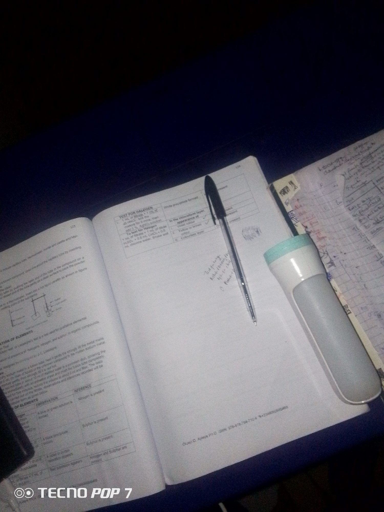 The image shows an open textbook on a dark blue surface, likely a table, under low light conditions. A black ballpoint pen lies on the left page of the textbook, and a white and blue LED flashlight is placed on the right page, partially obscuring some text and a notebook. The textbook has pages with scientific content, including tables and diagrams, and appears to be a chemistry or science textbook. Some handwritten notes are visible on the right page, as well as on a separate notebook placed to the right of the textbook. The lighting suggests it might be nighttime or a dimly lit room. The bottom left corner of the image features a circular logo with "TECNO POP 7" written inside. Based on the presence of the "TECNO POP 7" logo, which is a smartphone brand, and the general environment, this scene is likely captured in Nigeria, specifically in the city of Ipetumodu. The objects and their arrangement suggest someone is studying or working with the textbook and notes.