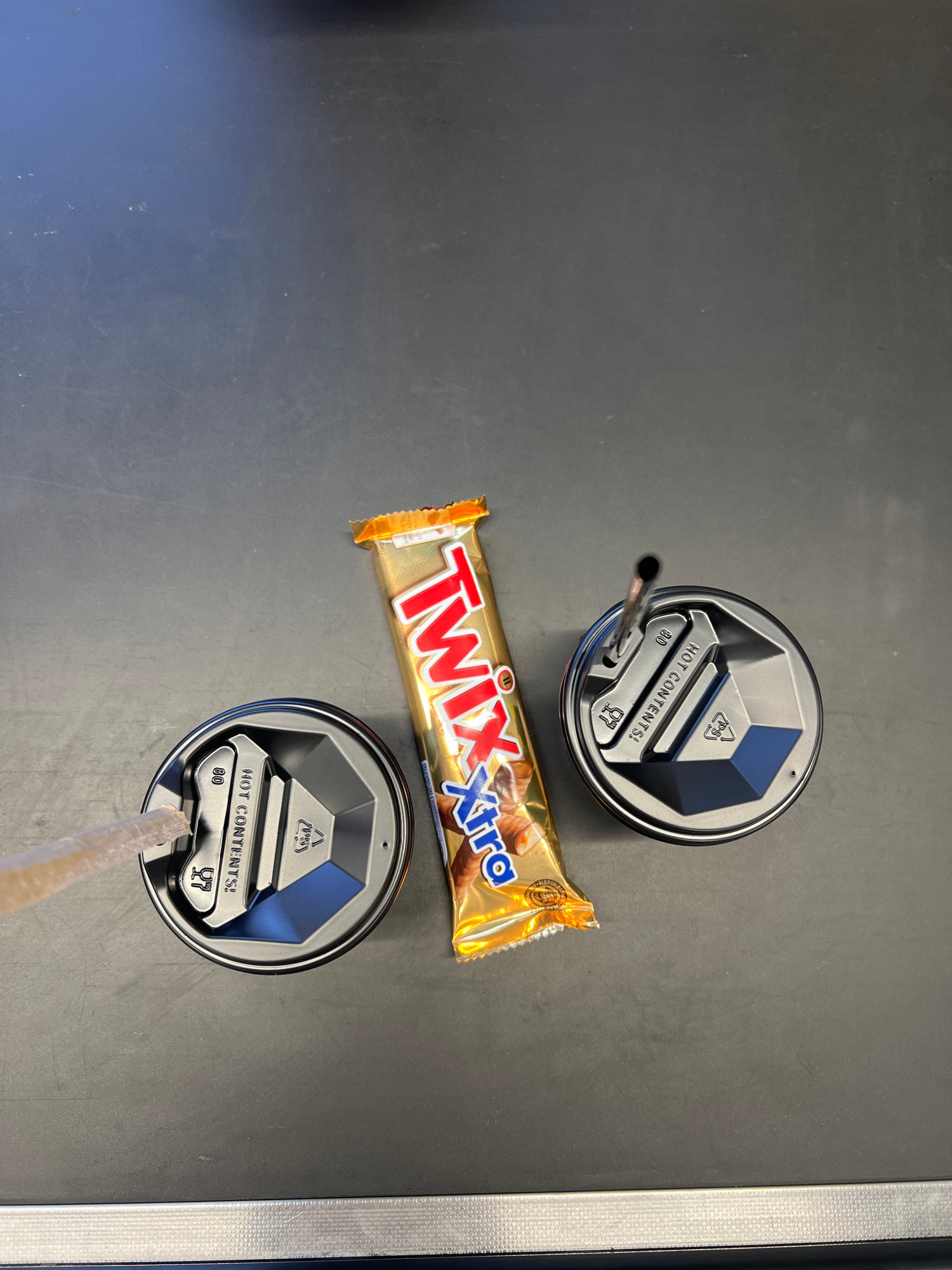 The image shows a Twix Xtra candy bar placed on a dark, flat surface, likely a table or countertop. To the left and right of the candy bar are two metallic travel mug lids, each with a straw or sipper mechanism. The lid on the left has a straw inserted, partially visible. The lid on the right has a straw inserted that extends upwards and is black. Both lids have embossed text reading "HOT CONTENTS!" and recycling symbols. The surface is textured and appears to be indoors. No people are visible in the image. The scene is not specific to any particular location in Kropyvnytskyi, Ukraine, as it lacks any identifying landmarks or context.