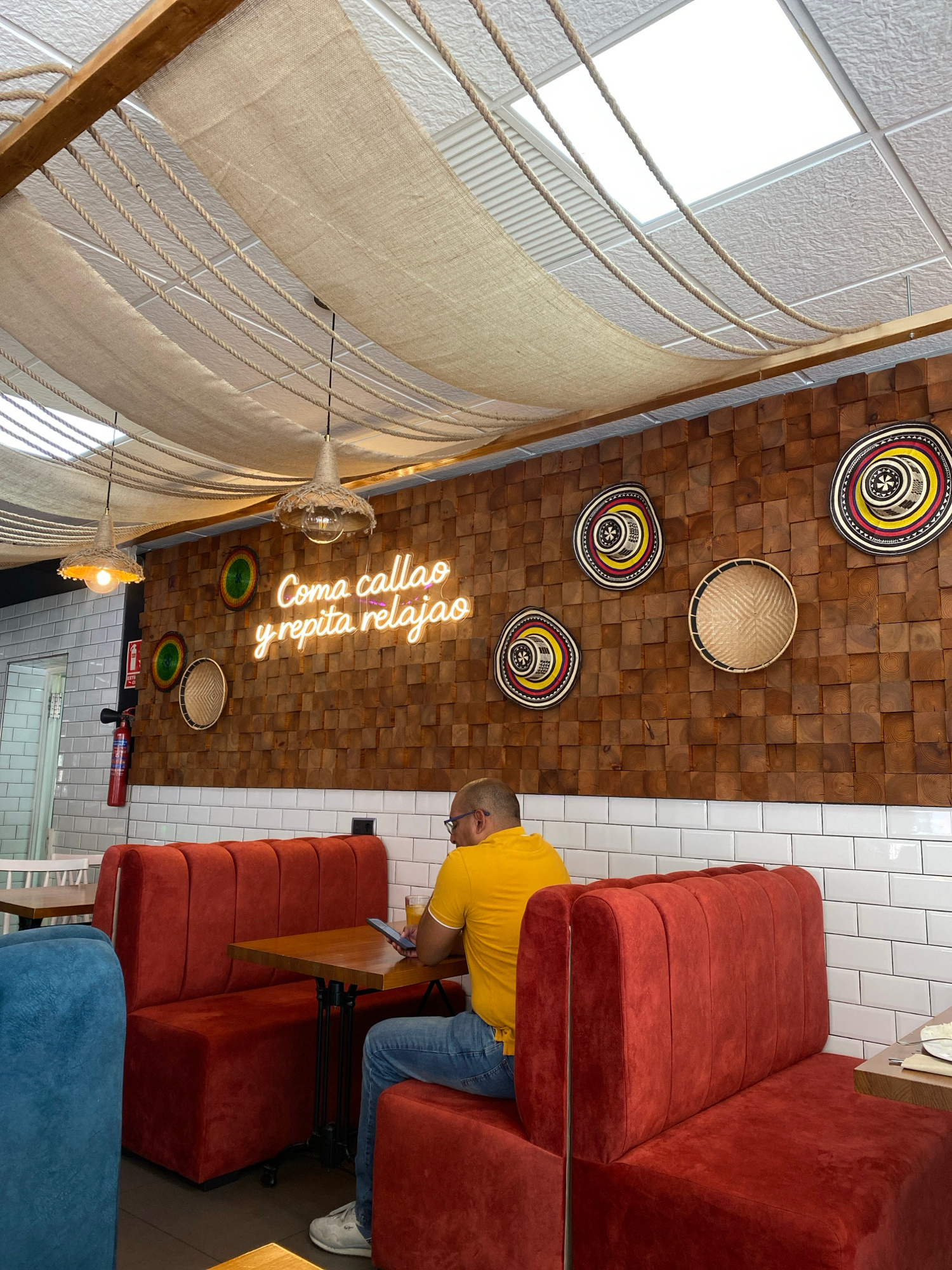 The image depicts the interior of a restaurant or cafe with a rustic and vibrant aesthetic. The ceiling is a prominent feature, constructed from a grid of wooden blocks and decorated with several traditional, colorful woven hats and baskets. A neon sign on the ceiling reads "Coma callao y repita relajo", which translates to "Eat and repeat relaxation".
In the foreground, a person wearing a yellow t-shirt and blue jeans is seated on a plush, rust-colored banquette, looking at their phone. This seating area faces a wooden table. Adjacent to the rust-colored seating, a bright blue banquette is partially visible. The walls are covered in white subway tiles.
The left side of the image reveals a distinct design element: a large, curved structure made of a rough, beige fabric draped over wooden beams and held by thick ropes. This evokes a nautical or tent-like feel. Two decorative lamps hang from this structure, featuring a woven shade.
The scene appears to be indoors, likely during the daytime given the bright natural light entering from what seems to be a skylight or large window at the top left of the image. The overall atmosphere suggests a relaxed and perhaps festive environment. There are no explicit weather cues visible. The location context provided is Murcia, Spain.