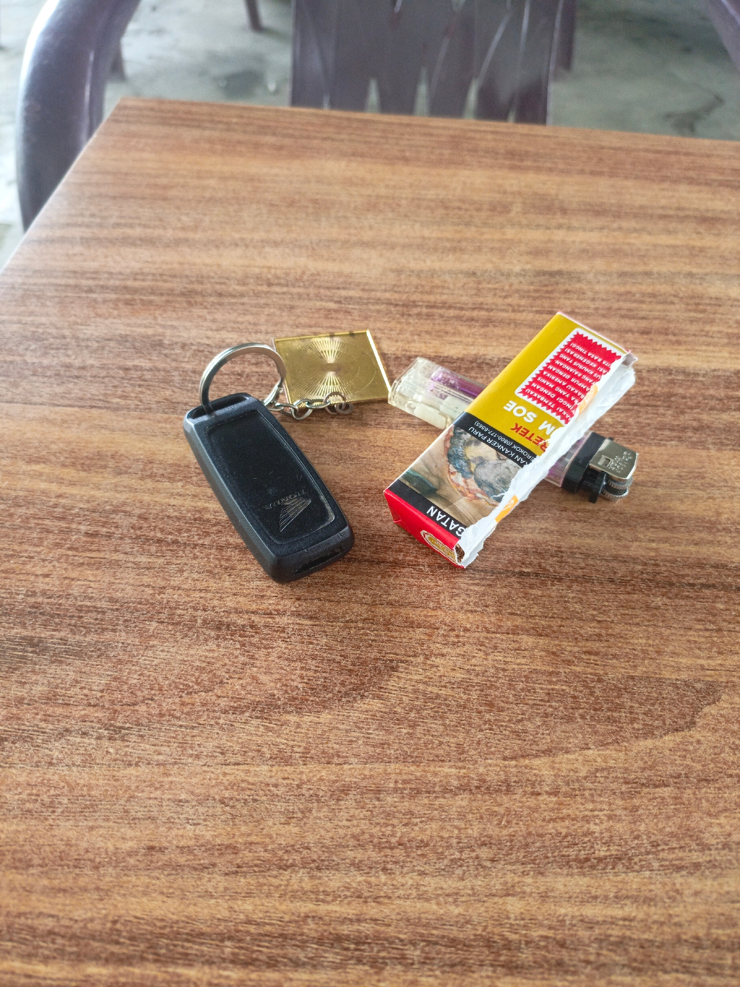 A close-up, eye-level shot shows a wooden table surface with several objects. In the foreground, on the left, is a black Honda key fob attached to a keyring with a small gold colored rectangular tag. To the right of the key fob is a crumpled cigarette pack, predominantly yellow with red and white text. The pack is partially covering a clear and purple disposable lighter. The background shows blurred chairs and what appears to be a concrete floor, suggesting an indoor or covered outdoor seating area, possibly in Lhokseumawe, Indonesia.