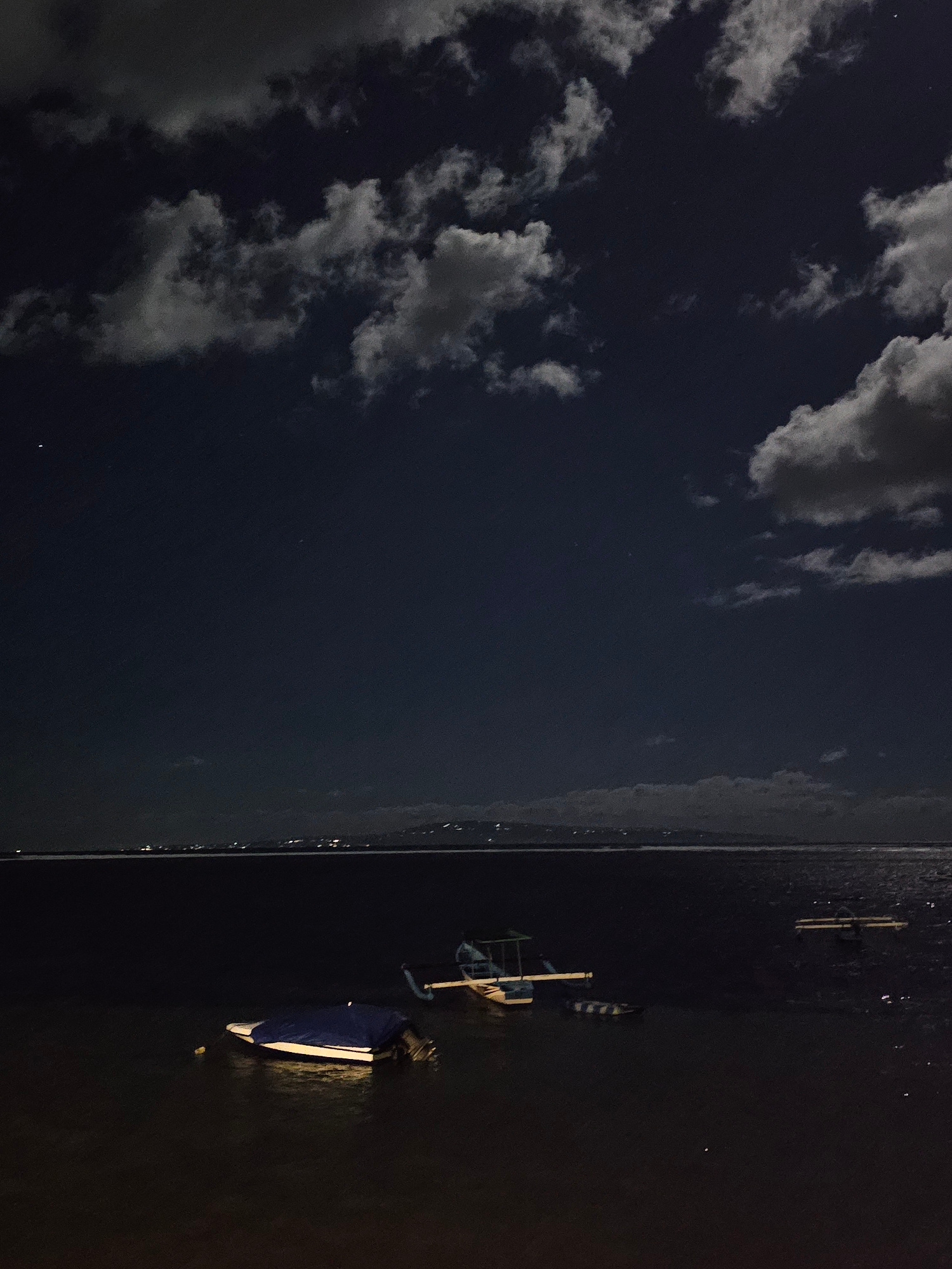 This image depicts a nighttime scene at a body of water, likely the coast of Denpasar, Indonesia. The sky is dark with scattered clouds, some of which are illuminated by moonlight, creating a textured, almost ethereal atmosphere. Several boats are visible on the water. In the foreground and slightly to the left, a small boat is docked, covered with a blue tarp, with its outboard motor submerged. Further out, a larger, lighter-colored boat with a canopy structure is partially visible. In the distance, the faint lights of a city skyline are discernible along the horizon, suggesting the location is near an urban area. The water appears dark and still, reflecting the dim light from the sky and the distant city. No people are visible in the frame.