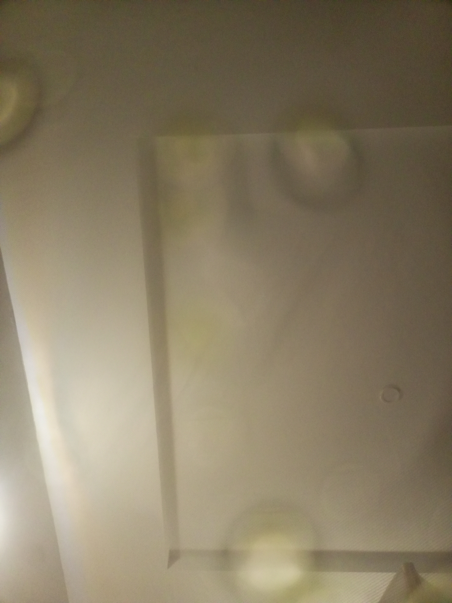 The image shows a ceiling with a recessed area and some subtle lighting effects or reflections. The ceiling appears to be a uniform off-white or light beige color. There is a distinct rectangular recess in the ceiling, with a visible edge. The rest of the ceiling is flat. There are some circular or oval shapes that appear to be light reflections or possibly water stains on the surface of the ceiling, particularly in the recessed area and towards the top left of the image. One small circular fixture, likely a light or smoke detector, is visible on the right side of the flat ceiling. The overall lighting in the scene is dim and diffused. The image is taken from a low angle, looking upwards towards the ceiling. The location is Unknown, Unknown.