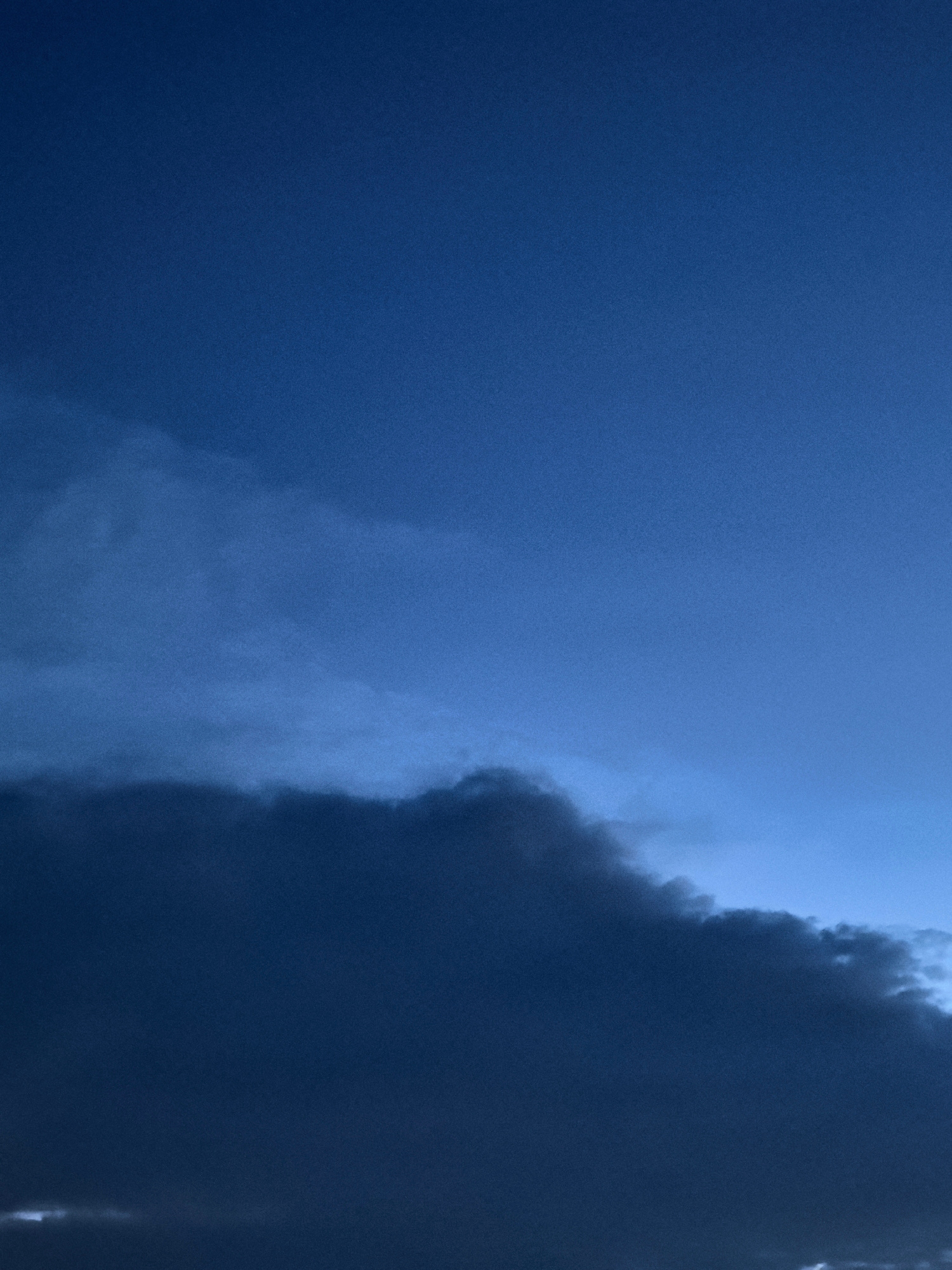 A low-angle, full shot captures a dark, ominous cloud formation against a deep blue twilight sky. The clouds, dense and heavy with rain, dominate the lower half of the frame, obscuring the horizon. A faint, lighter cloud layer is visible above the darker mass, suggesting layers of atmospheric activity. The sky above is a gradient of deep blue, transitioning to a lighter shade towards the top of the image. No other details are visible.