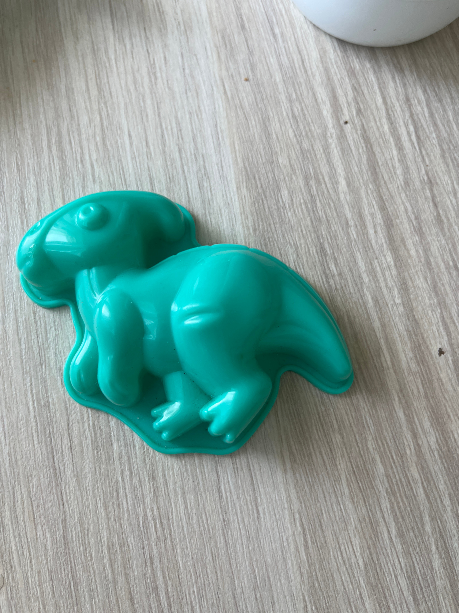 A teal-colored plastic dinosaur mold is placed on a light-colored, wood-grained surface. The mold is in the shape of a Parasaurolophus, with its distinctive crest visible. The dinosaur is depicted in a crouching pose, with its front legs slightly bent and its tail curving to the right. The surface beneath the mold has a texture of parallel lines, suggesting it is a wooden tabletop or floor. No people or other objects are visible in the immediate vicinity of the mold. There is no information available about the location in Chelyabinsk, Russia.