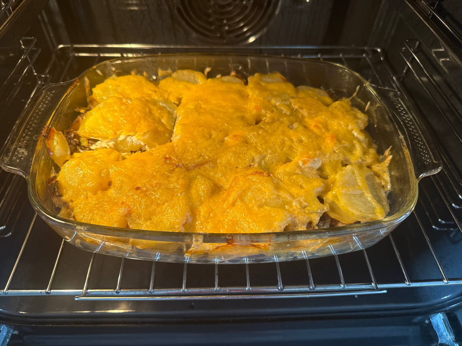 A close-up shot captures a glass baking dish filled with a cheesy potato and meat casserole, positioned on a metal rack inside an open oven. The casserole is golden brown on top, with melted cheese covering layers of sliced potatoes and what appears to be ground meat. The oven's interior, including the heating element and rack, is visible. The lighting suggests it might be daytime, possibly during cooking hours, but no specific time can be determined. The setting is a domestic kitchen, likely in Tolyatti, Russia, given the context. The image focuses on the food being prepared, implying a mealtime event or food preparation activity. There is no visible text in the image.