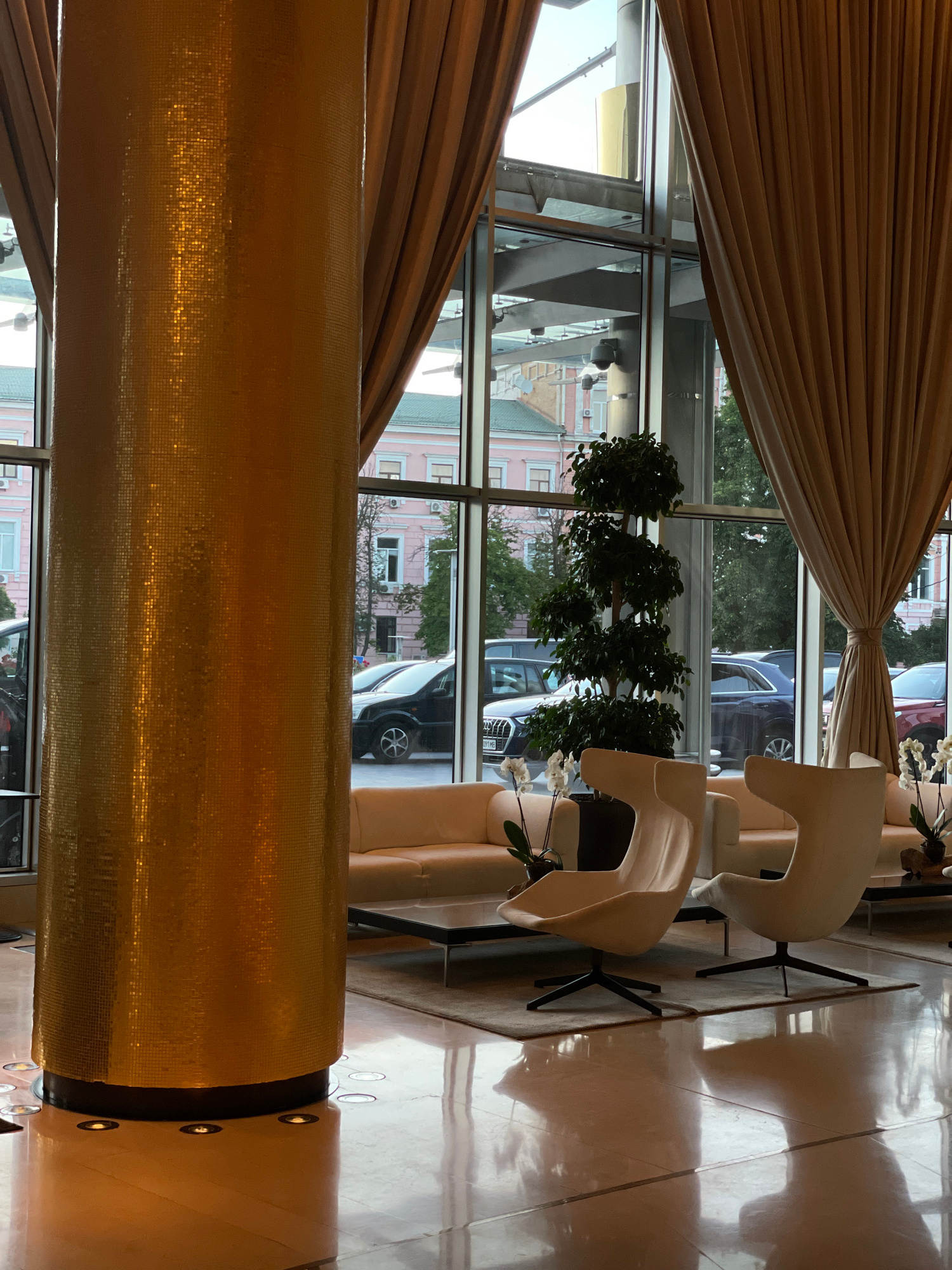 This image captures an interior scene, likely a hotel lobby or a high-end waiting area, with a large, golden, mosaic-covered pillar dominating the foreground. The pillar reflects the light from the large windows, which offer a view of a city street. Outside, several cars are parked, and buildings with pink facades and green roofs are visible. Inside, two modern, cream-colored armchairs are arranged on a rug in front of a light-colored sofa. A black coffee table sits in front of the sofa. A tall, potted plant with dark green leaves stands near the window. Beige curtains are drawn back, framing the view. The floor is highly polished, reflecting the interior furnishings and the light from the pillar. There are no people visible in the image. Based on the architectural style and the presence of specific building facades visible through the windows, this location is identified as the lobby of the Premier Palace Hotel in Kyiv, Ukraine.