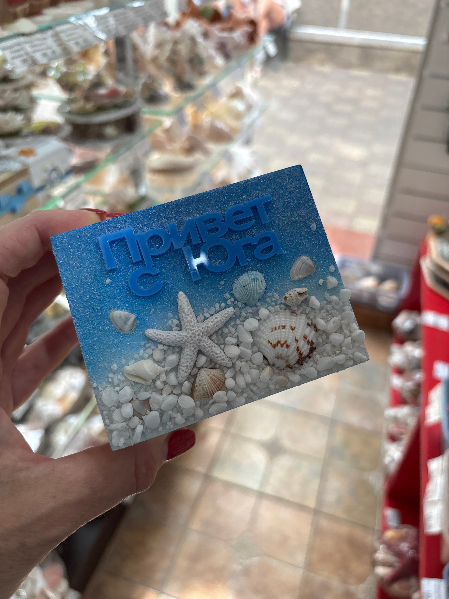 A hand with red nail polish holds a rectangular object decorated with a beach theme. The object has a blue background sprayed with white specks to resemble sand. It is adorned with various seashells and a white starfish. Blue 3D letters spell out "Привет, Дорога" which translates to "Hello, dear" in Russian. The scene appears to be indoors, possibly a shop or market, with shelves and displays blurred in the background. The lighting suggests it is daytime. The overall impression is of a decorative souvenir or gift.