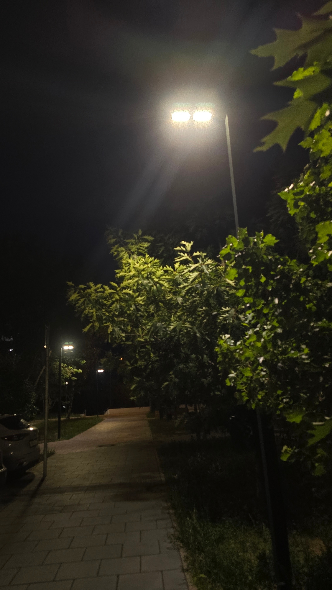 A wide, low-angle shot captures an outdoor scene at night. A bright, rectangular street lamp illuminates the upper right portion of the frame, casting light rays into the dark sky. To the left of the lamp, a tall, thin pole extends upwards. In the foreground, a paved walkway made of light-colored rectangular tiles runs diagonally from the bottom left corner towards the upper right. Trees with vibrant green leaves are prominent in the mid-ground and right side of the image. Some leaves are illuminated by the street lamp, appearing a lighter, almost yellow-green. In the background, a dark, paved path is visible, also illuminated by streetlights, suggesting an urban or park setting. A white car is parked on the left side of the walkway. The overall atmosphere is one of a quiet, well-lit urban pathway at night.