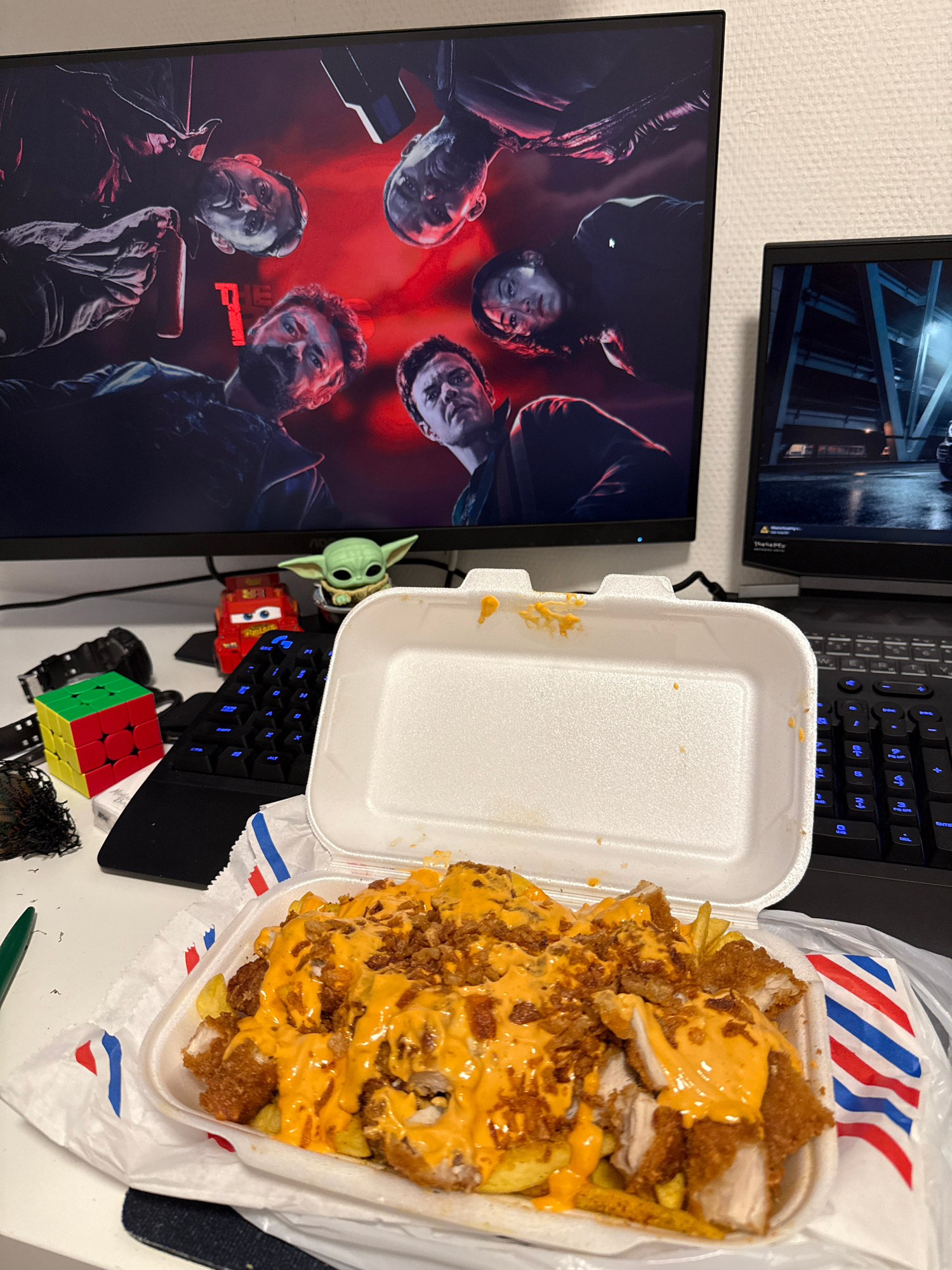 The image shows a desk setup with two computer monitors, a keyboard, and a white styrofoam food container. The left monitor displays a poster for "The Boys" TV series, featuring several actors' faces. The right monitor shows a scene from a video game, possibly a car driving through a dimly lit urban environment. In the foreground, the food container holds a meal consisting of fried chicken pieces and fries, covered in an orange sauce and topped with crispy fried onions. Various desk accessories are visible, including a small Rubik's Cube, a toy car resembling Lightning McQueen from the movie "Cars," and a figurine of Baby Yoda. A black watch is also present. The background appears to be a wall with a textured finish. The scene is indoors, likely a personal workspace or entertainment area. There is no indication of the city of Enschede, Netherlands.