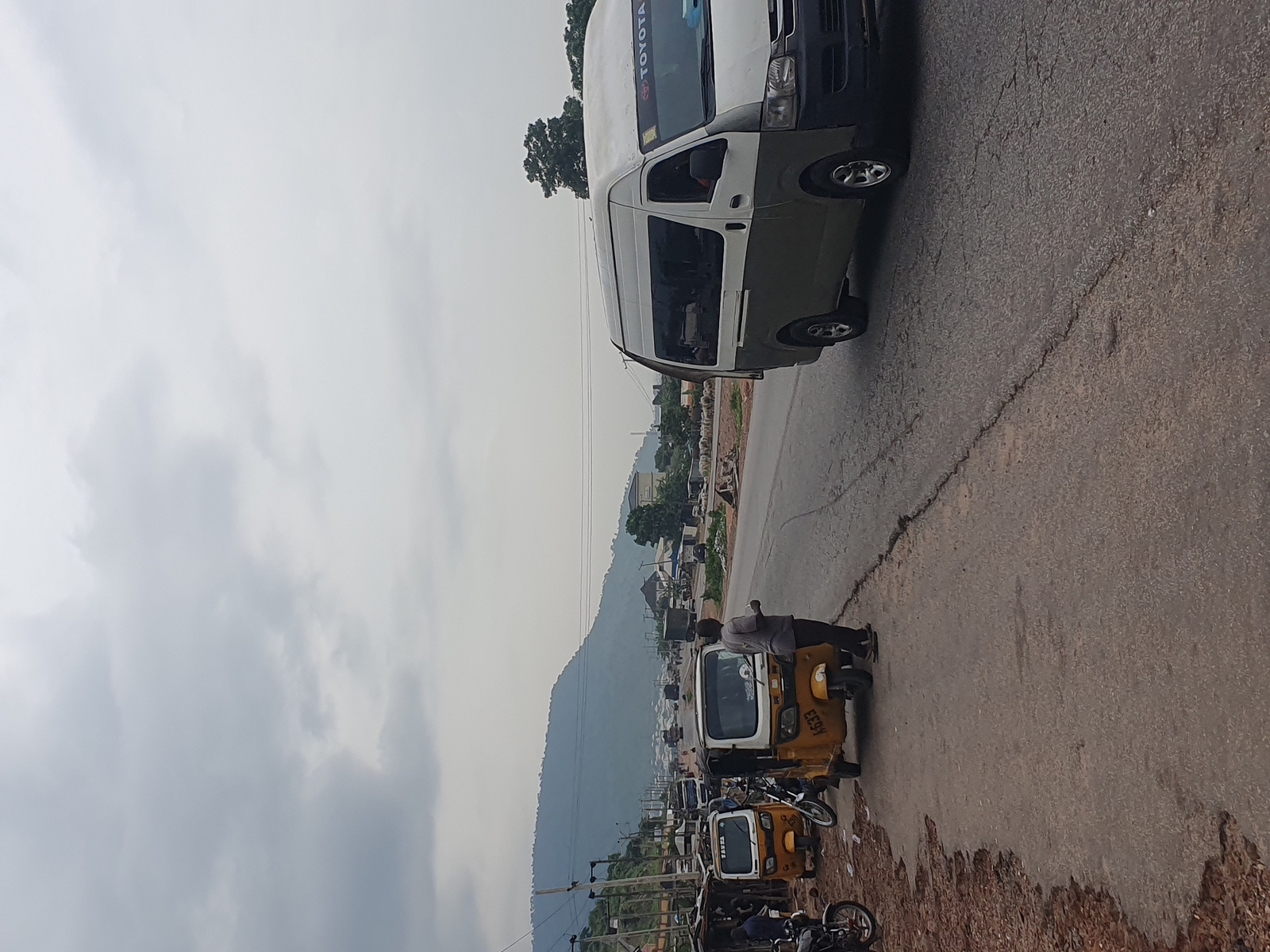 A wide-angle shot taken from a slightly elevated perspective captures a busy street scene in Lokoja, Nigeria, under an overcast sky. The image is oriented vertically. In the foreground, the cracked asphalt road takes up a significant portion of the right side. A yellow auto-rickshaw, known locally as a "Keke Napep," is parked on the right side of the road. A man is standing near the auto-rickshaw, appearing to be a passenger or driver. Further down the road, another similar auto-rickshaw and several motorcycles are visible. A white and green minibus, identified by a "TOYOTA" sticker on its windshield, is parked on the left side of the road, facing away from the viewer. Trees and vegetation are scattered along the left side of the road, with a mountainous backdrop dominating the scene in the distance. Buildings are clustered along the road, indicating a developed area within the city. The overall impression is one of a bustling commercial or transportation hub in Lokoja.