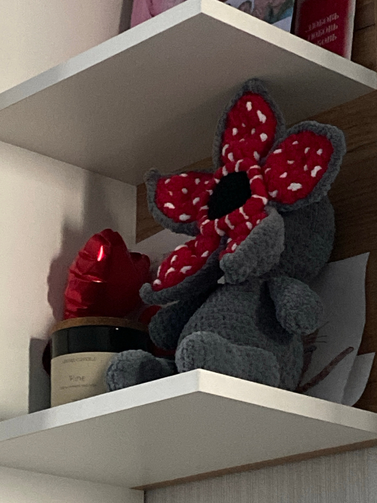 The image shows a stuffed toy, possibly a Demogorgon from Stranger Things, sitting on a white shelf. To the left of the toy is a red, vaguely flower-shaped object with a glowing red light in the center. Below the red object and to the left of the toy is a jar with a black lid and a label that reads "Pine." The background behind the shelf appears to be a wooden surface. The image is taken from a slightly elevated angle, looking down at the objects on the shelf. There is no indication of the location being Chelyabinsk, Russia.