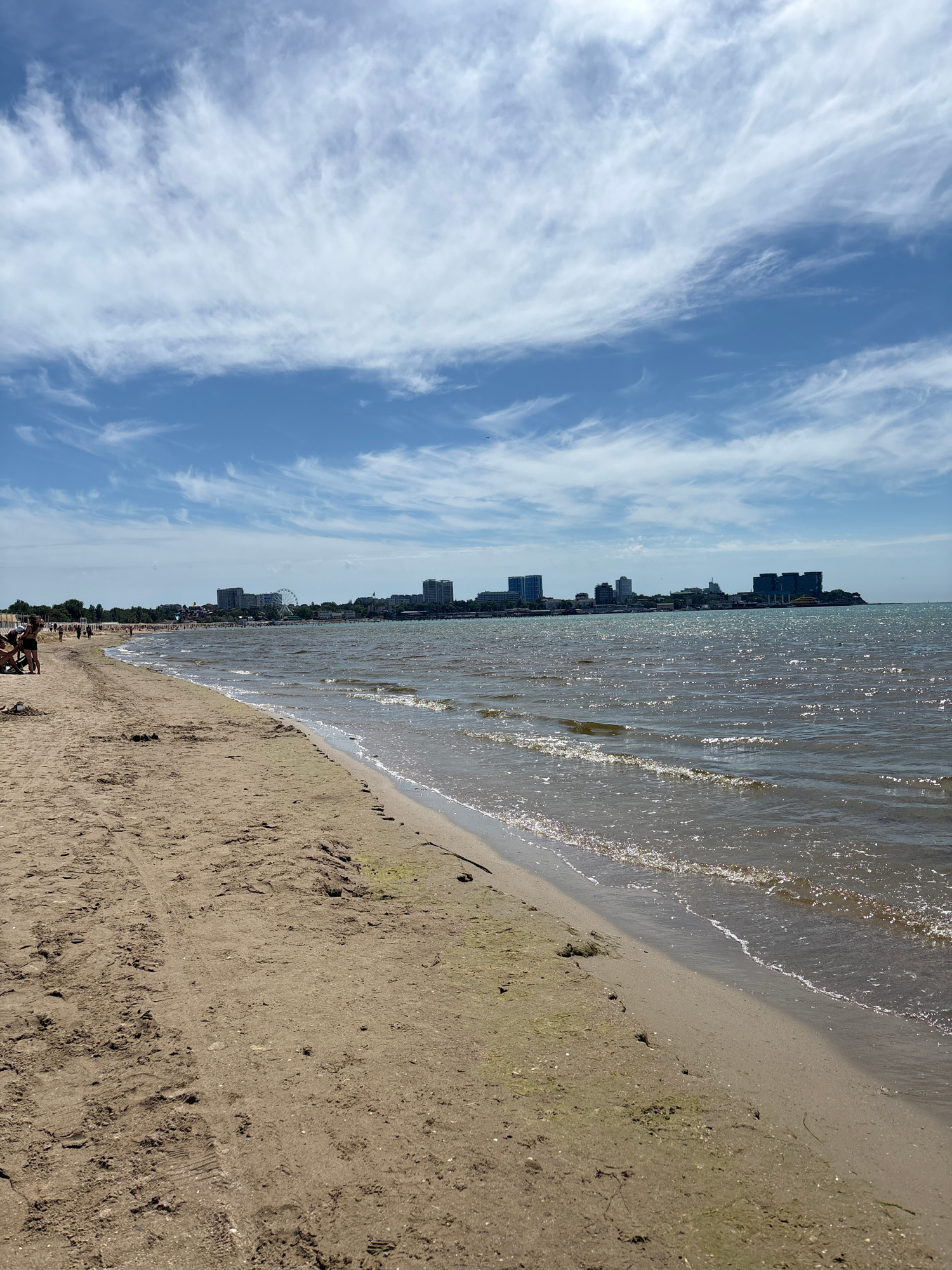 This image captures a vibrant daytime scene at a beach in Anapa, Russia. The composition features a wide expanse of bright blue sky adorned with long, wispy white clouds, suggesting fair weather. Below the sky, the sparkling sea stretches across the mid-ground, its surface reflecting the sunlight in numerous glints. Gentle waves break along the sandy shoreline, creating a clear demarcation between the water and the beach.
The foreground and right side of the image are dominated by the sandy beach. The sand appears light brown and somewhat damp closer to the water, with subtle texture and a few footprints visible. Scattered along the beach in the mid-ground are several indistinct figures, suggesting people relaxing or walking, enjoying the coastal environment.
Along the horizon, a distant city skyline is visible, characterized by numerous multi-story buildings, likely hotels or apartment complexes, lining the coast. A notable landmark, a Ferris wheel, stands out among these structures, indicating a recreational area. The overall impression is one of a popular resort town enjoying a sunny day. No visible text is present in the image.